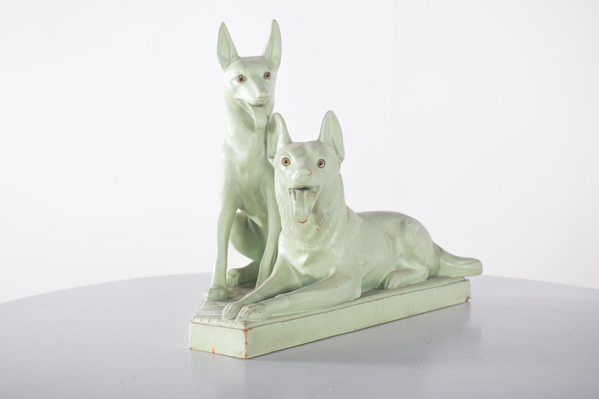 Scultura in argilla dipinta che raffigura due cani. Decorato in verde pastello, raffigura un cane sdraiato con la bocca leggermente aperta, mentre l'altro è seduto con le orecchie erette e un'espressione attenta. Entrambi hanno occhi con dettagli
