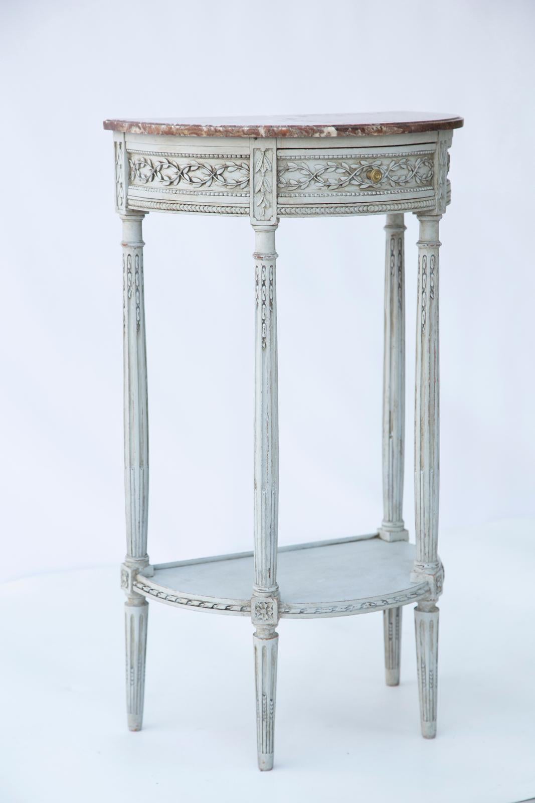 Demilune hall table, having a top of Breccia Vino marble, on conforming table base painted in light blue showing natural wear; its fielded apron carved with intertwined foliate vines, centered by a single frieze drawer, raised on round, stop-fluted,