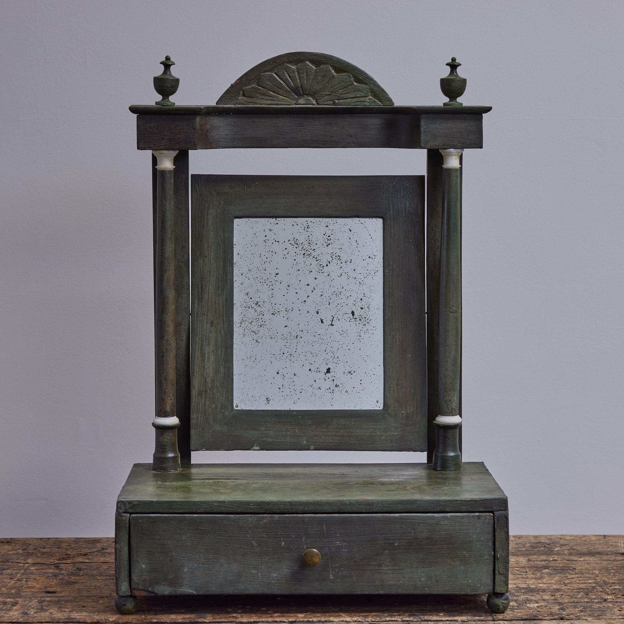 Green painted dresser mirror from 1840s England. The tilt mirror is flanked by two ionic columns and a floating frieze with carved fan pediment and urn finials. A rectangular base rests on ball feet and contains a drawer with a simple round brass