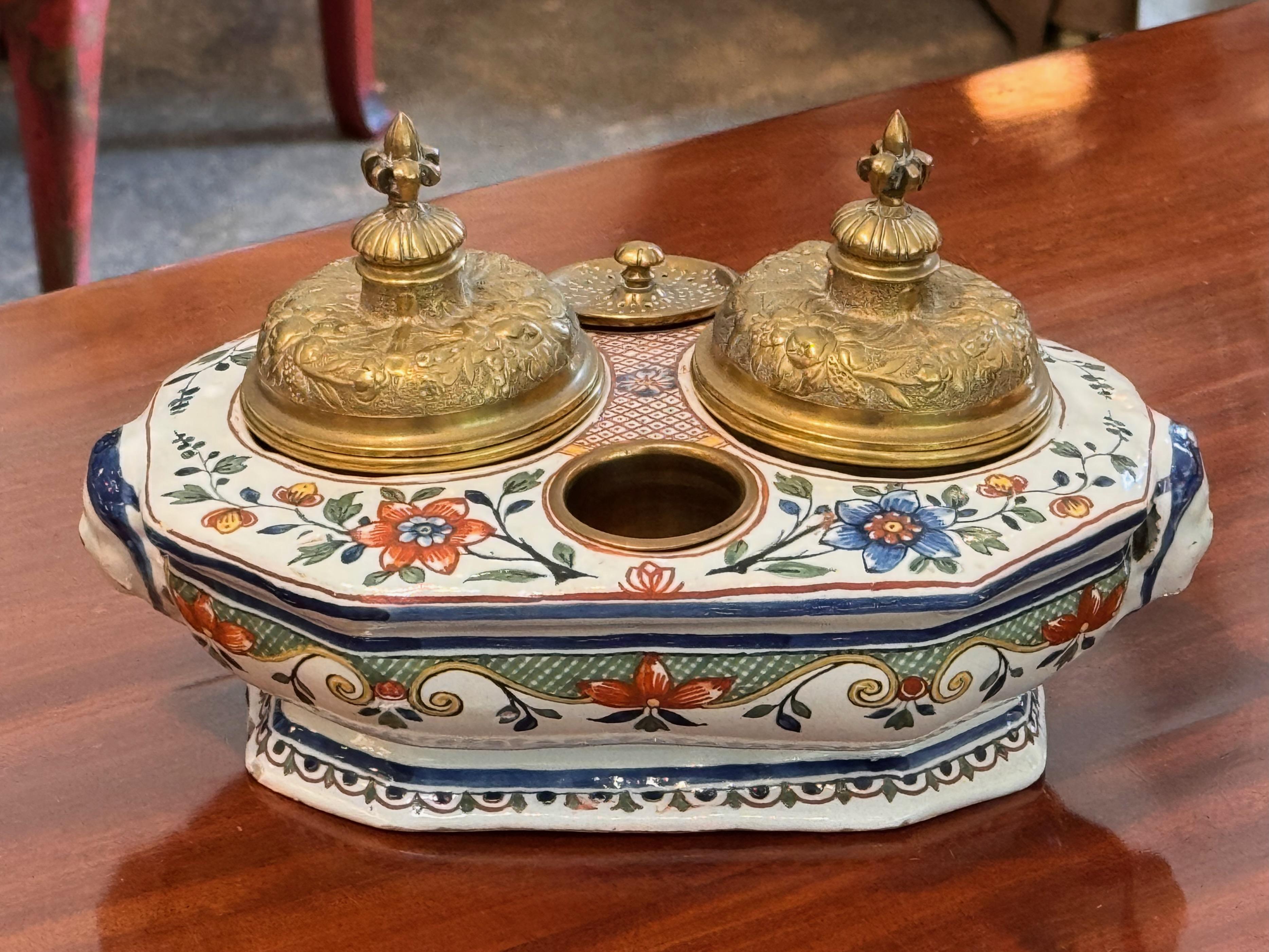 Tintero de colorida loza con motivos florales pintados a mano en rojo, verde y azul, con tapas de latón dorado finamente detalladas y rematadas con flor de lis. El tintero incluye forros desmontables de latón y cristal, y un pocillo central