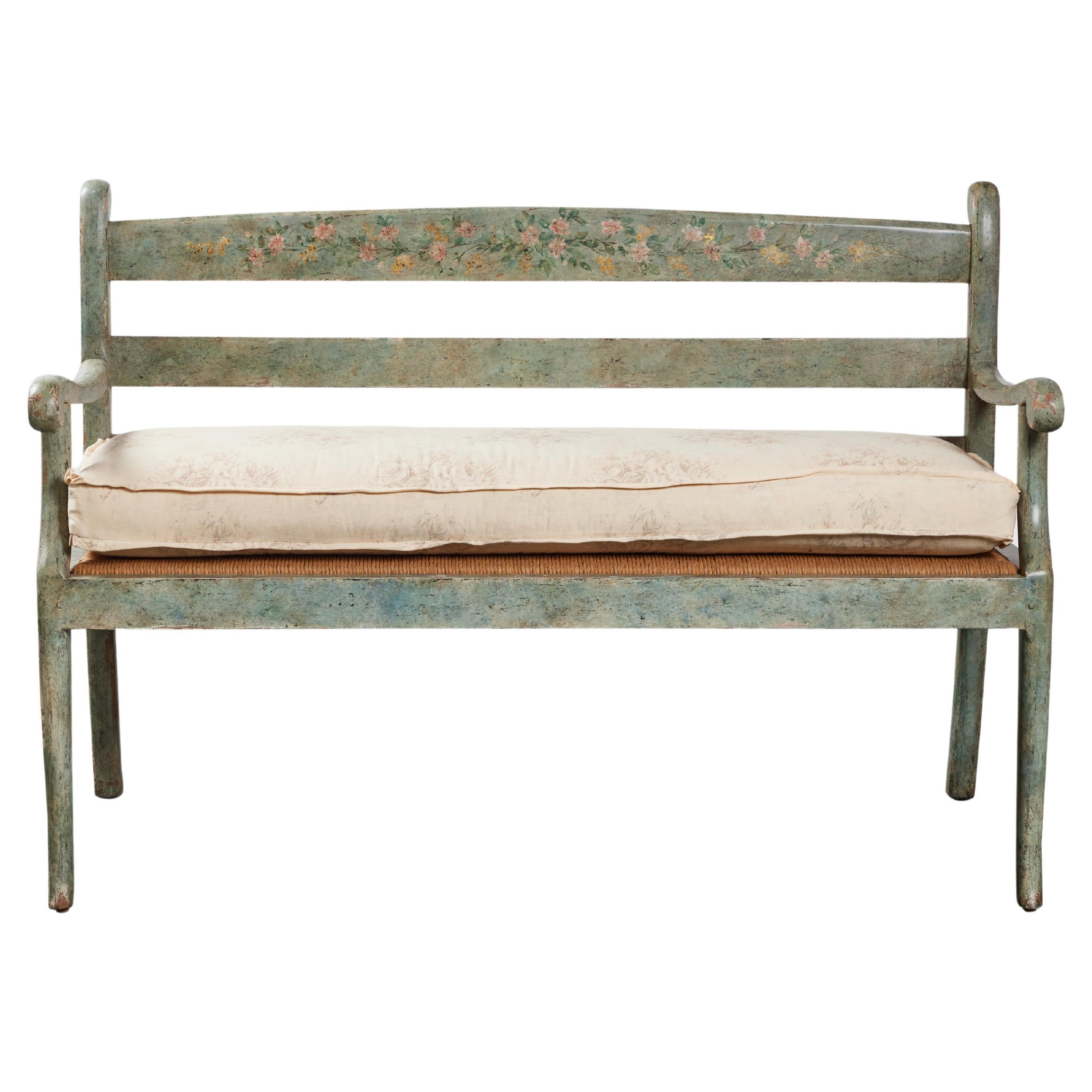 Painted French Provincial Style wooden bench with wicker seat and custom upholstered cushion. Original green paint with floral design on the backrest. Made by John Hall Designs, Los Angeles, 1990. 

Additional Dimensions: 
Arm Height 28