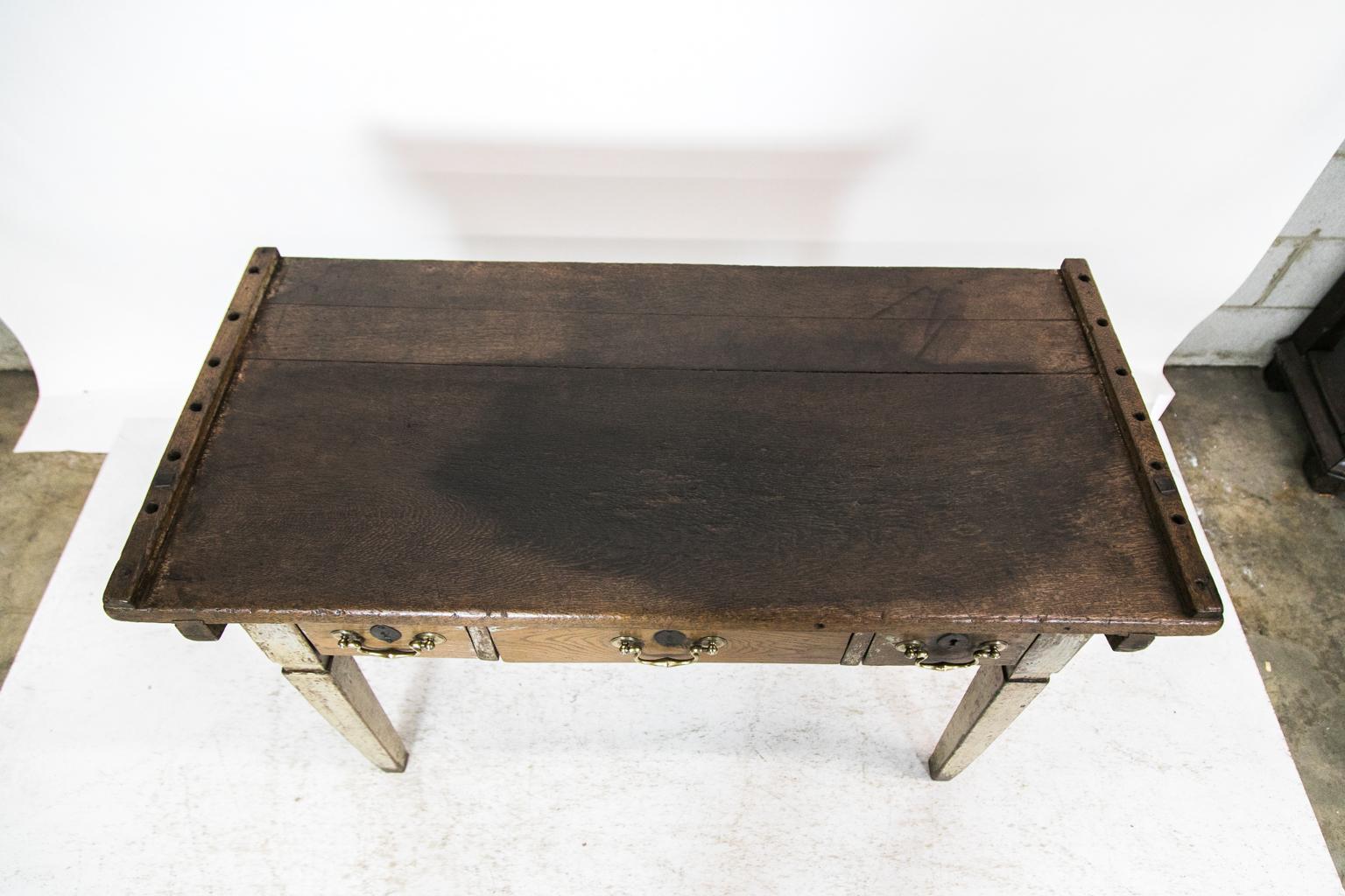 This work table has square tapered legs. There are traces of the original gray-blue paint throughout. The wood is oak and has a 1 1/8-inch-thick top which is secured to the base with 1 3/8-inch-thick dove tailed batten cleats that are secured with