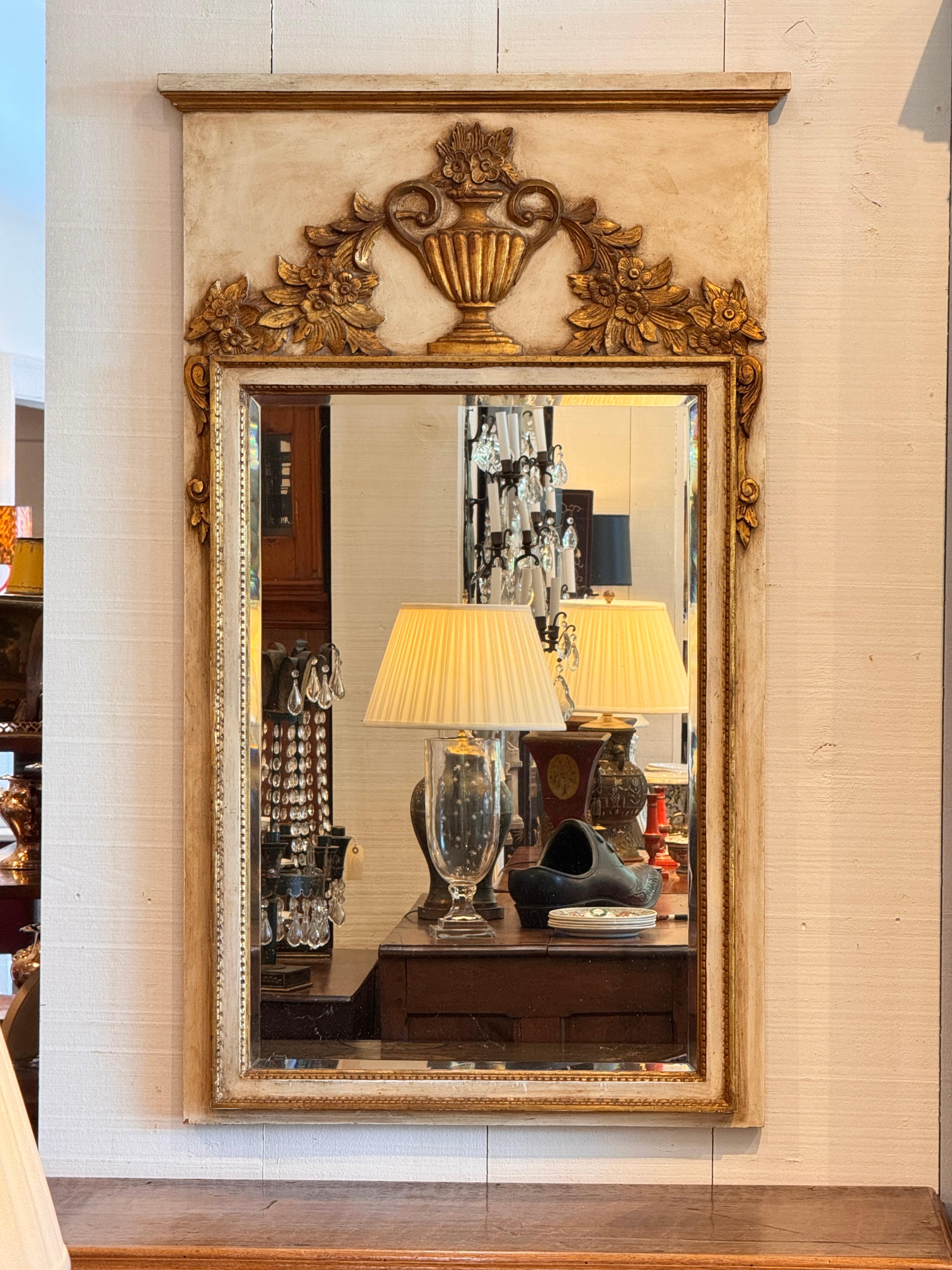 Miroir peint italien représentant une urne sculptée et dorée avec des guirlandes florales sur un cadre rectangulaire. L'entourage de couleur crème contraste avec les détails dorés, rehaussant ainsi le relief décoratif. Une garniture perlée borde la