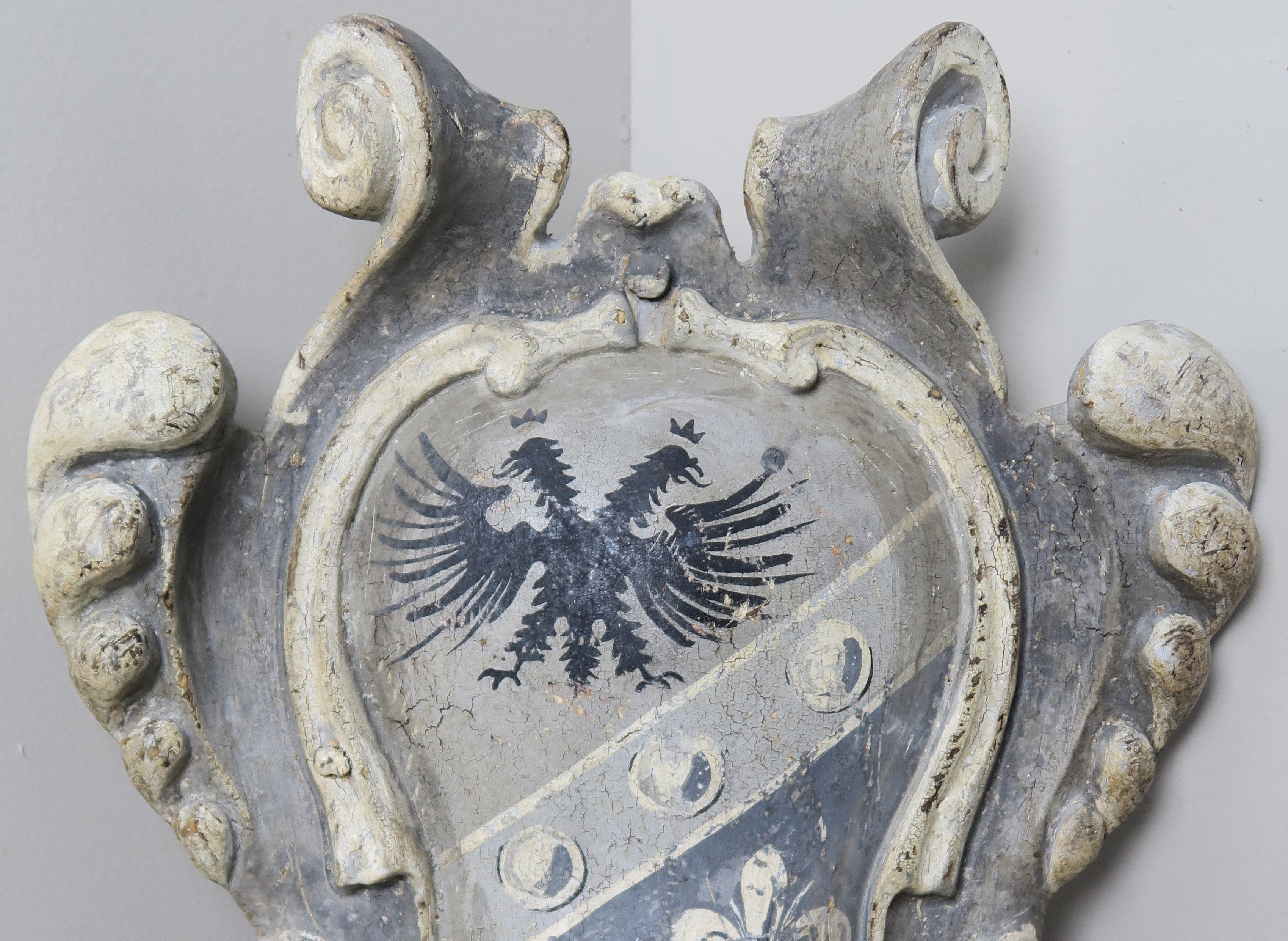 Italian wood scrolled shield depicting a two headed winged dragon and fleur de lys. The shield is hand painted in soft shades of gray and antique white.
