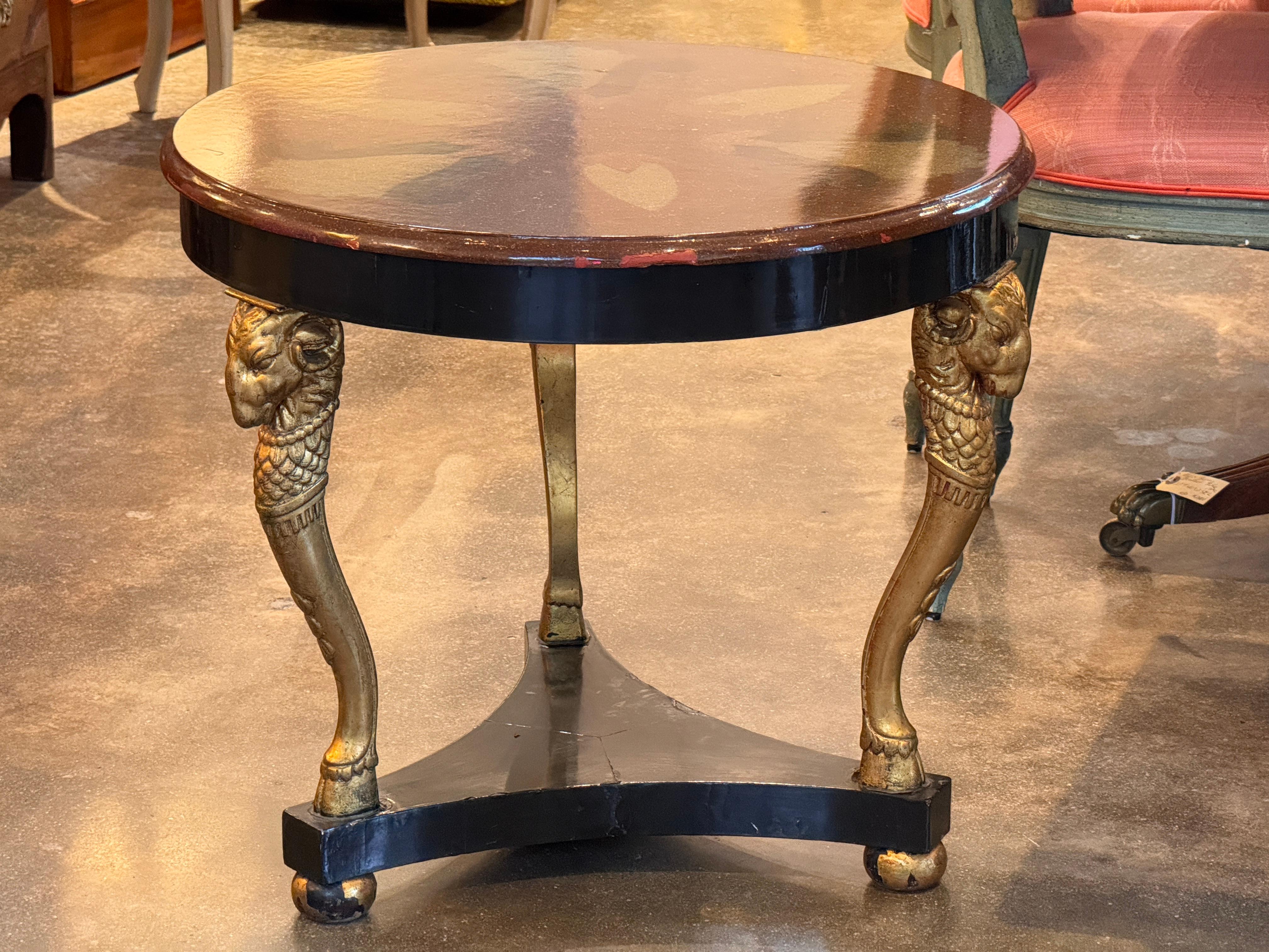 Bemalter runder niedriger Tisch mit einer dekorativen Platte aus Kunstmarmor und vergoldeten Widderkopfbeinen. Der schwarz gestrichene Sockel bildet eine dreieckige Streckbank, die die skulpturale Präsenz verstärkt. Die an der Schürze sichtbare