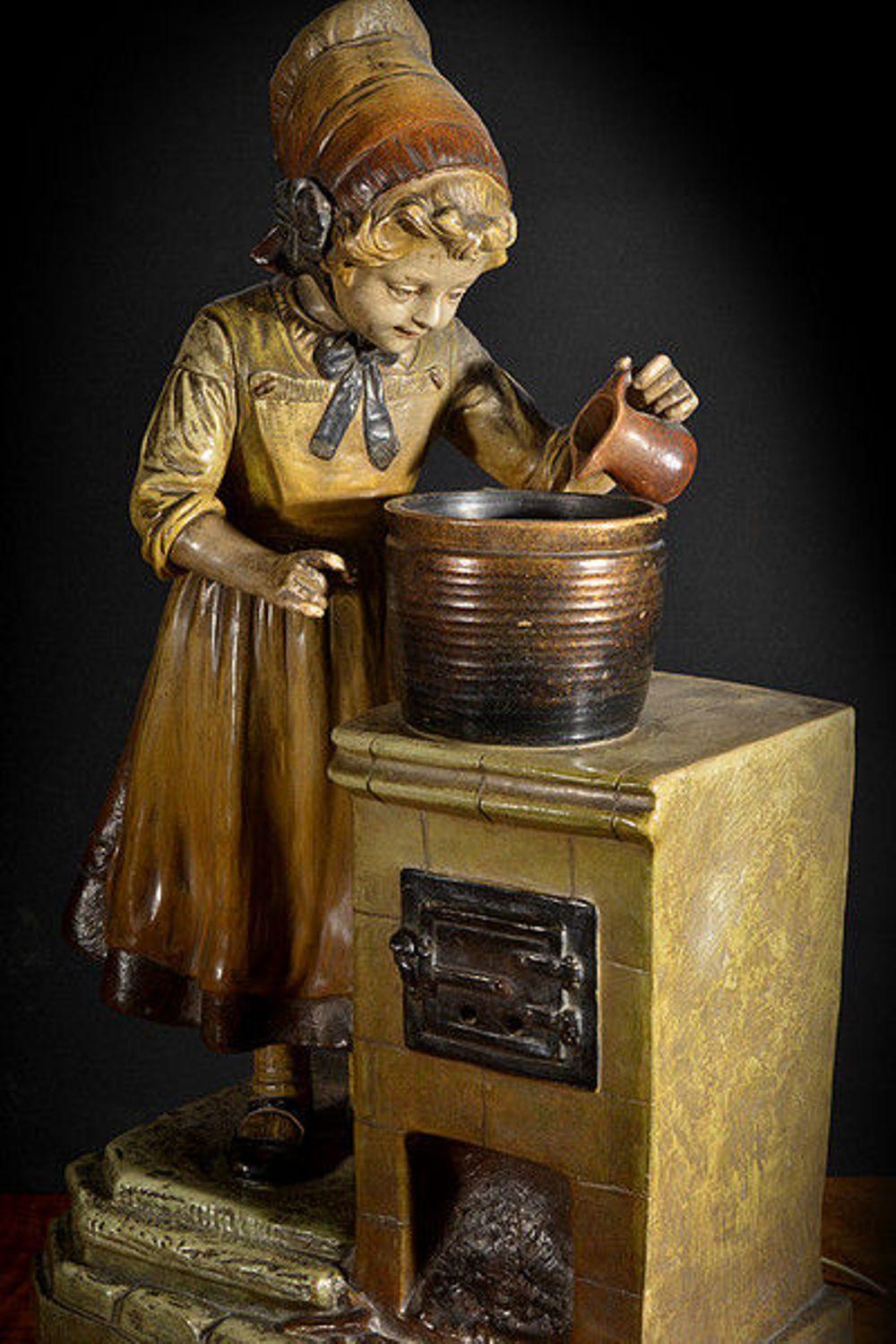 An unusual painted metal lamp depicting a young girl pouring the contents of a small jug into a large pot which sits on a stove.

The girl, wearing an apron over a dress, Mary Anne shoes and a bonnet with a ribbon tied to one side, looks happily