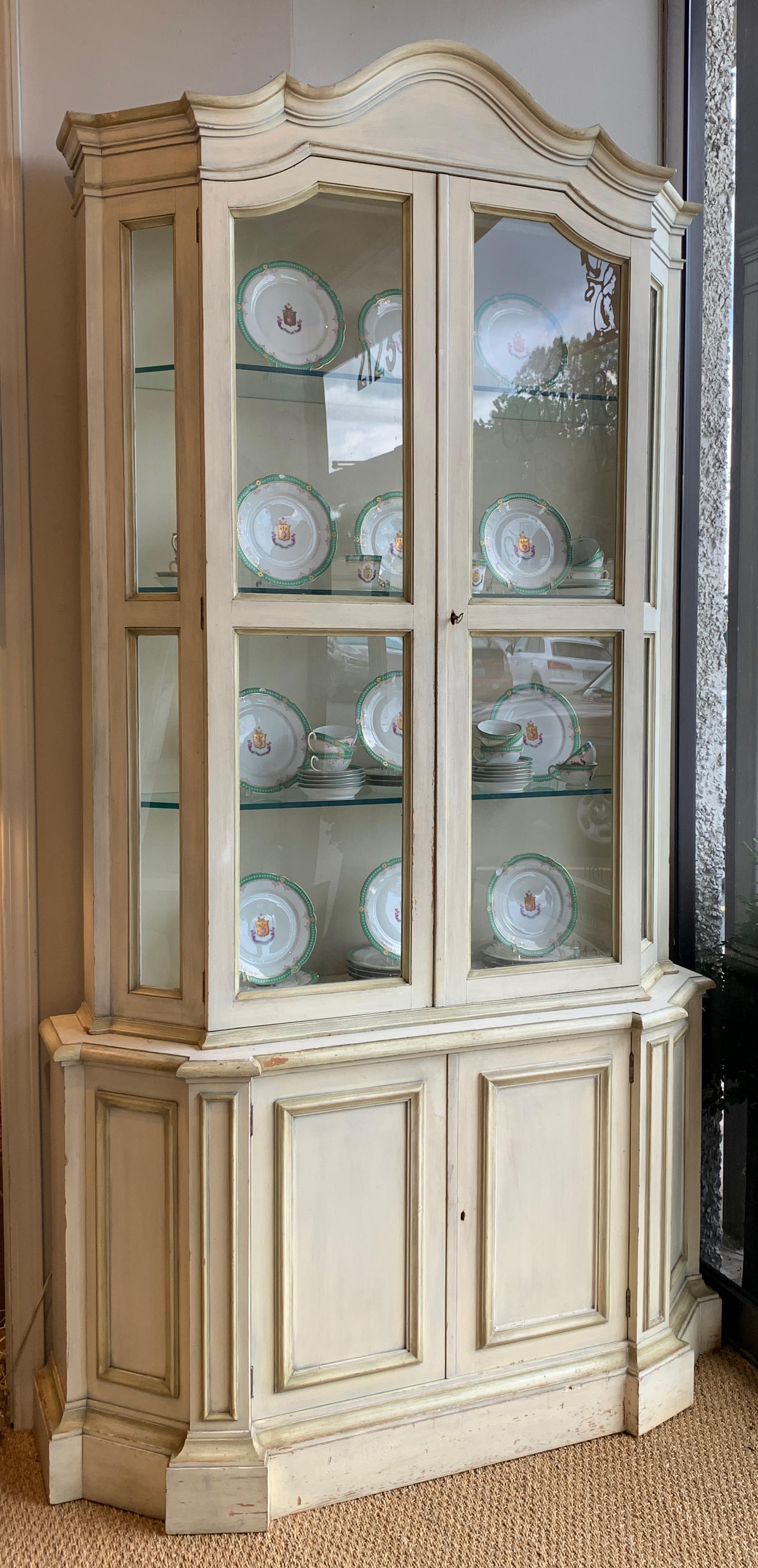 A tall and elegant mid-20th century. Paint decorated china or display cabinet with carved bonnet top and two glass doors revealing three substantial glass shelves above two cabinet doors offering additional storage.
