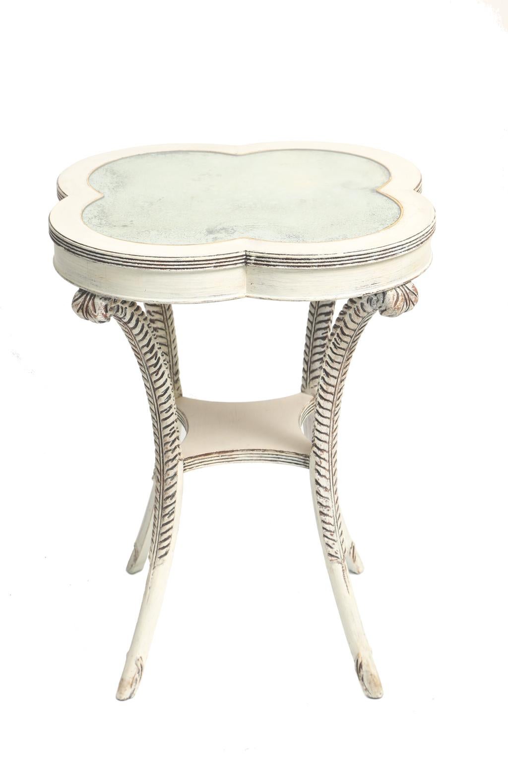 Table, having a painted finish showing natural wear, its quatrefoil top inset with antiqued mirror, raised on four, plume-carved, splayed legs, joined by concave shelf stretcher. 

Stock ID: D2120.