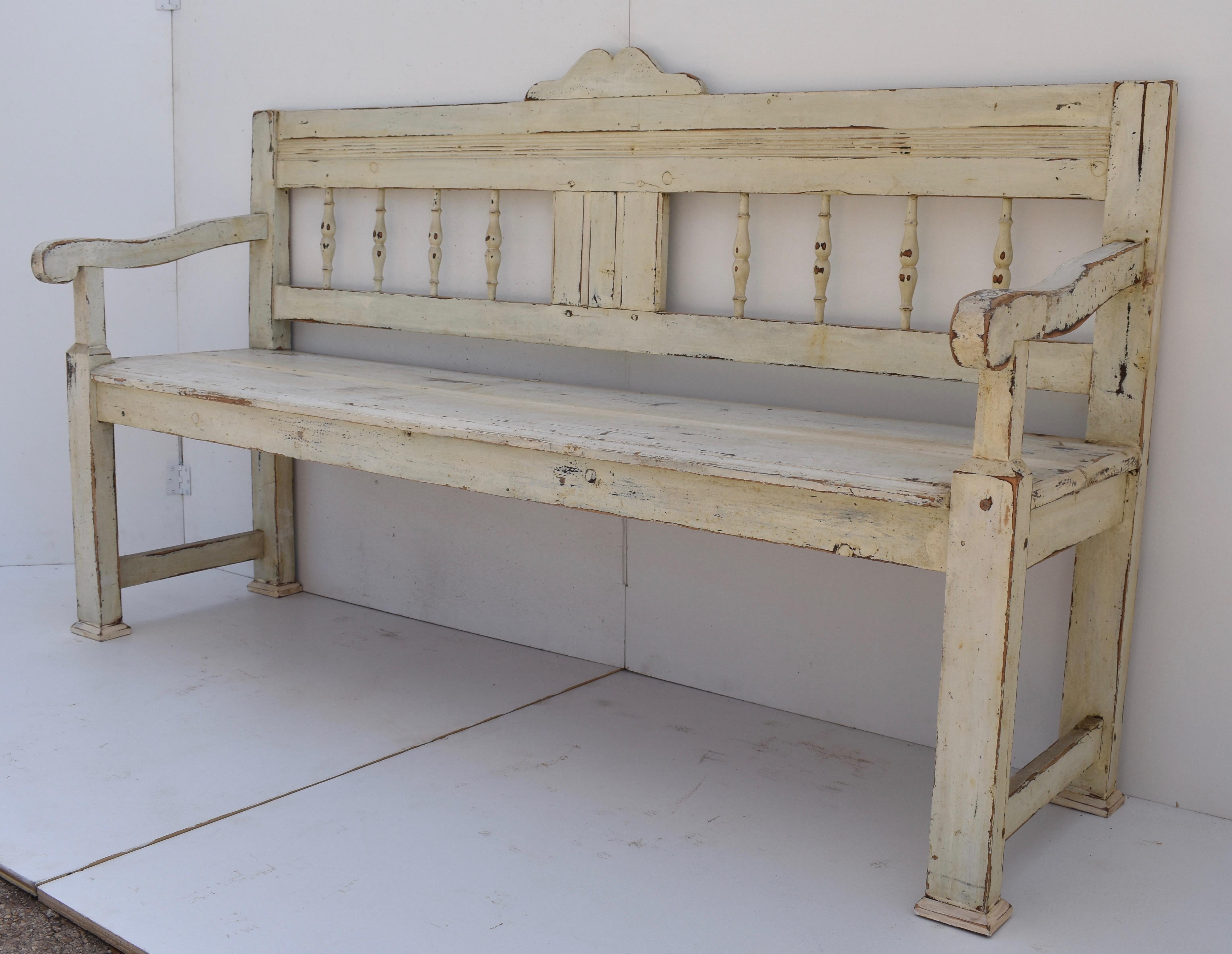 This rather stately pine bench has a small scallop cresting a bold top rail above a central faux paneled splat flanked by four turned spindles each side. The shapely protruding arms are supported by nicely tapered front legs. In old white paint worn