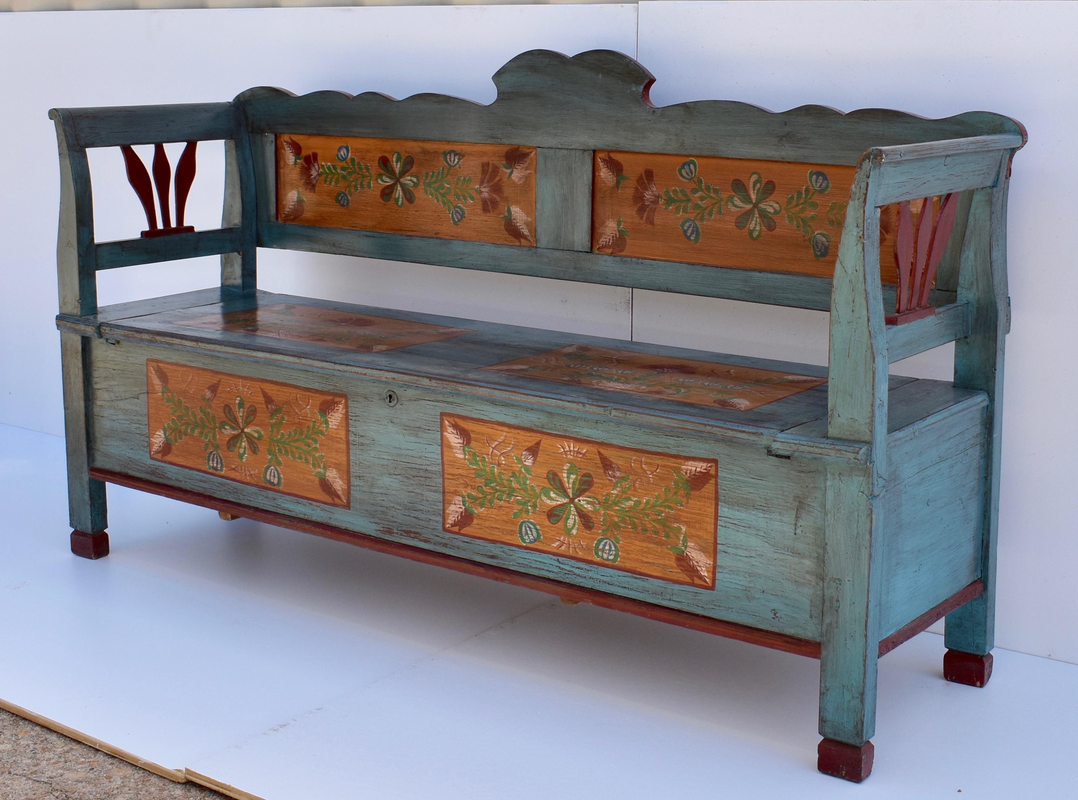 This handsome and stately antique pine storage bench has been recently painted by an accomplished Hungarian folk artist. The scalloped back rail rests above two flat panels. The integral legs and arms are gently scrolled at the top. The seat lifts