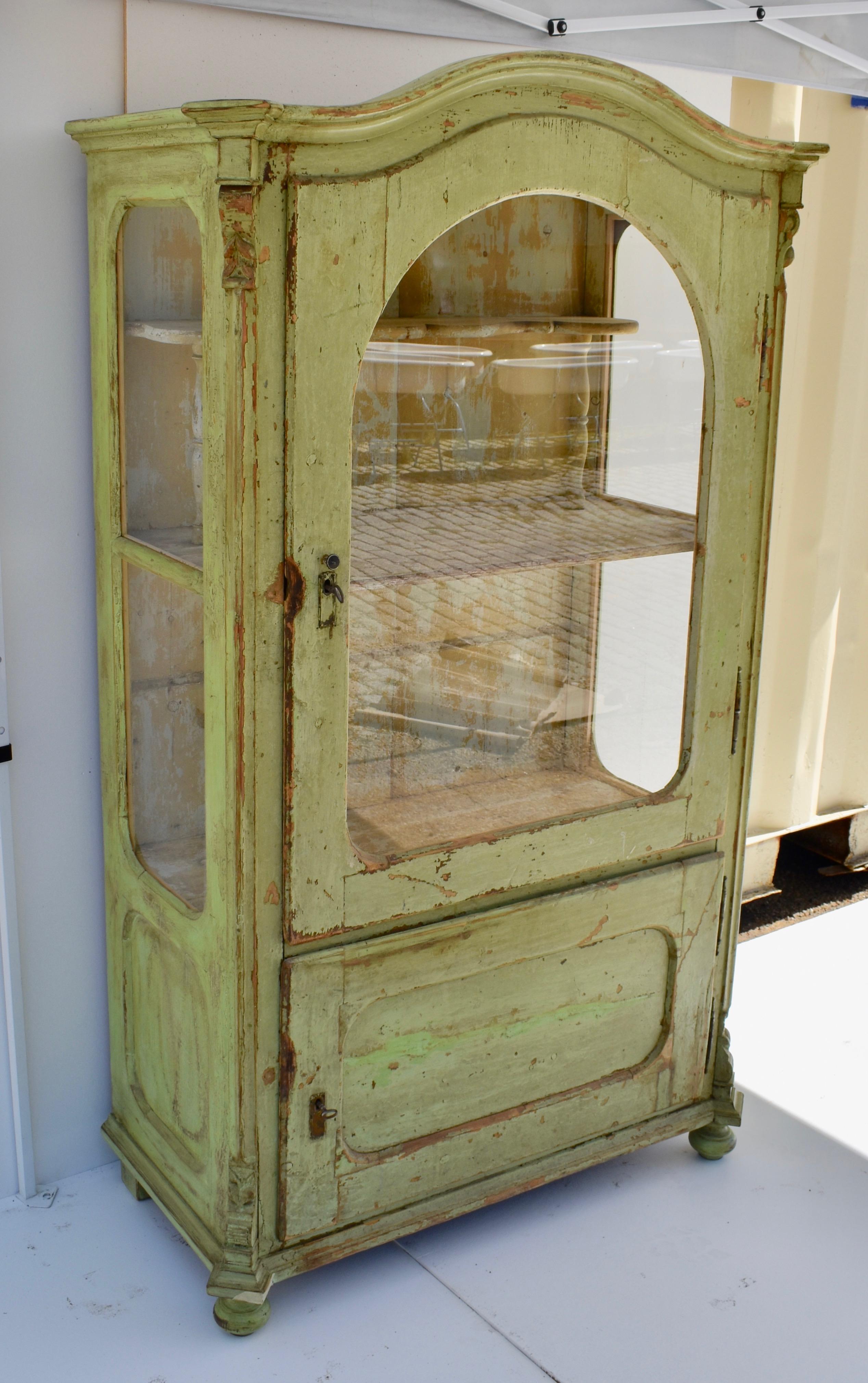 This interesting two door vitrine or glazed cabinet features a bonnet top above an arched glazed door with a flat paneled door beneath, both mounted on wide-swinging fiche hinges. The upper interior has a central full shelf with a decorative gallery