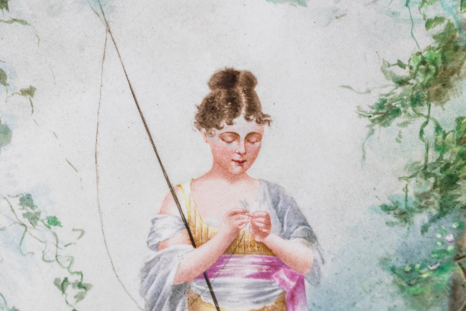 Painted porcelain plate figuring a fisherwoman near a lake in the middle of nature. She holds a fishing rod under her arm and tries to hung an insect on the hook. She wears a pink, orange and white dress and a white cloth on her shoulders. She wears