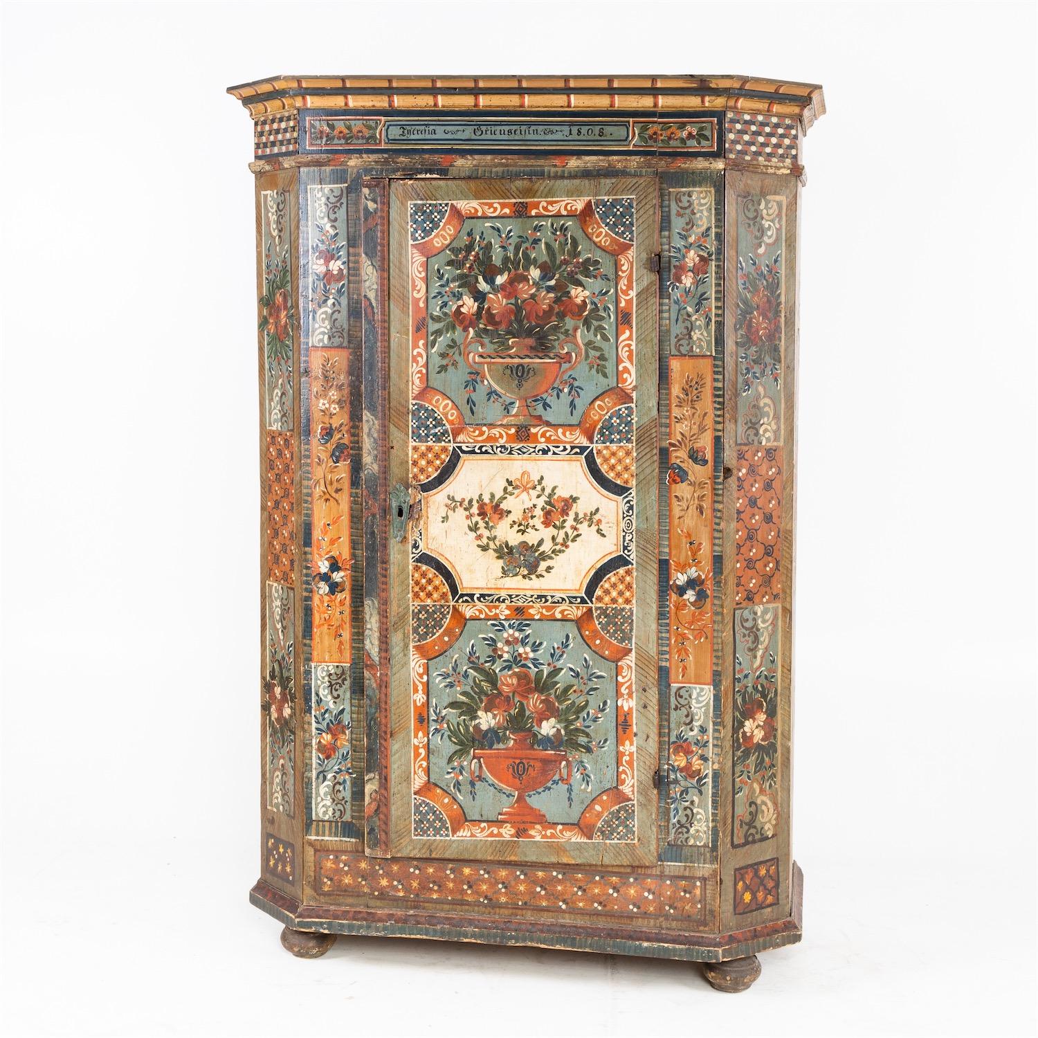 Provincial cupboard with beveled corners and profiled cornice standing on bun feet. The single-door body is painted on all sides and shows floral bouquets in paneled frames. The cornice bears the inscription in a cartouche field: 