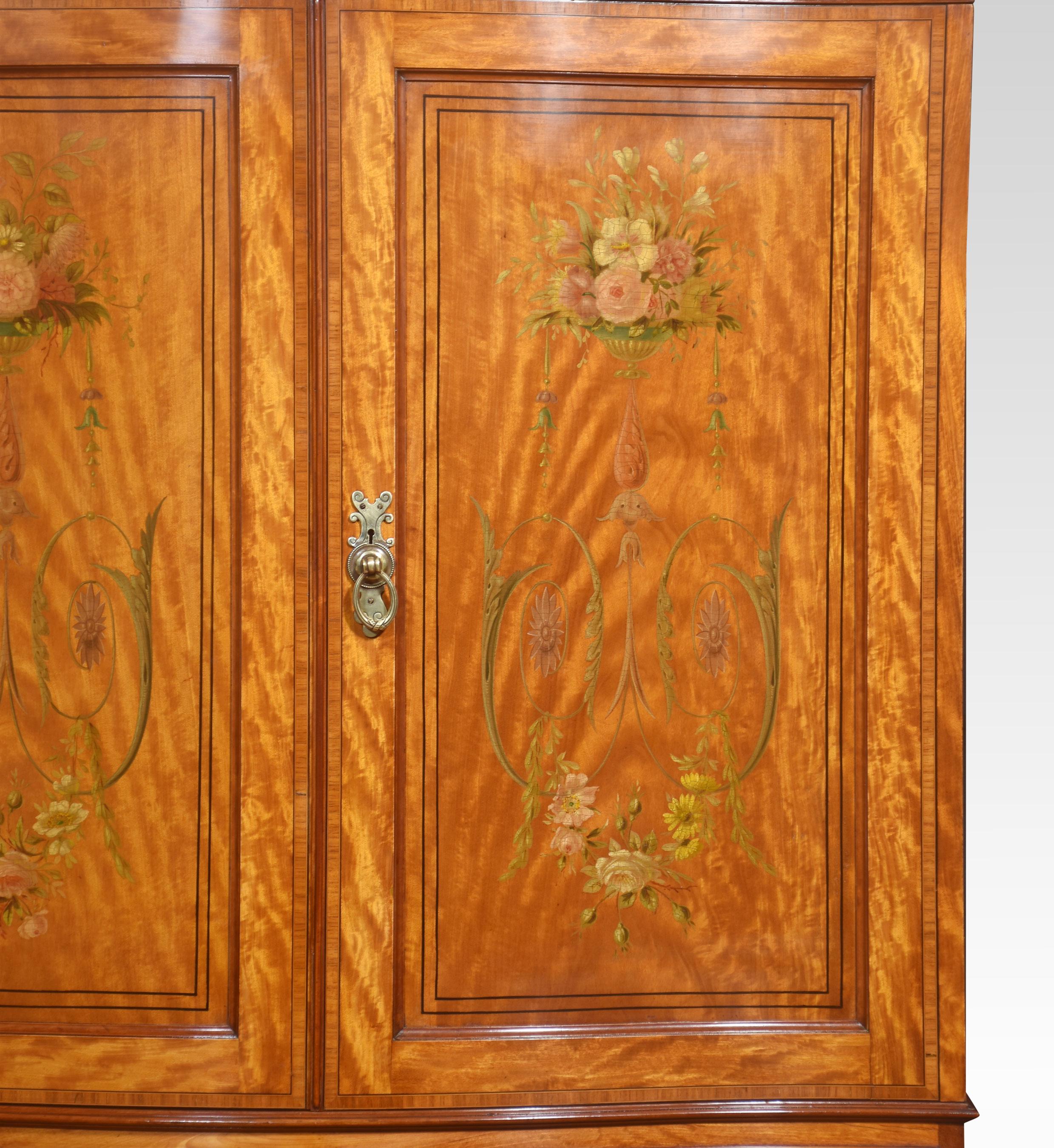 Satinwood wardrobe of serpentine outline, having carved moulded cornice above a pair of well-figured satinwood doors with floral painted decoration. The doors open to reveal a large hanging area which is utilised by having a faux drawer in the base.