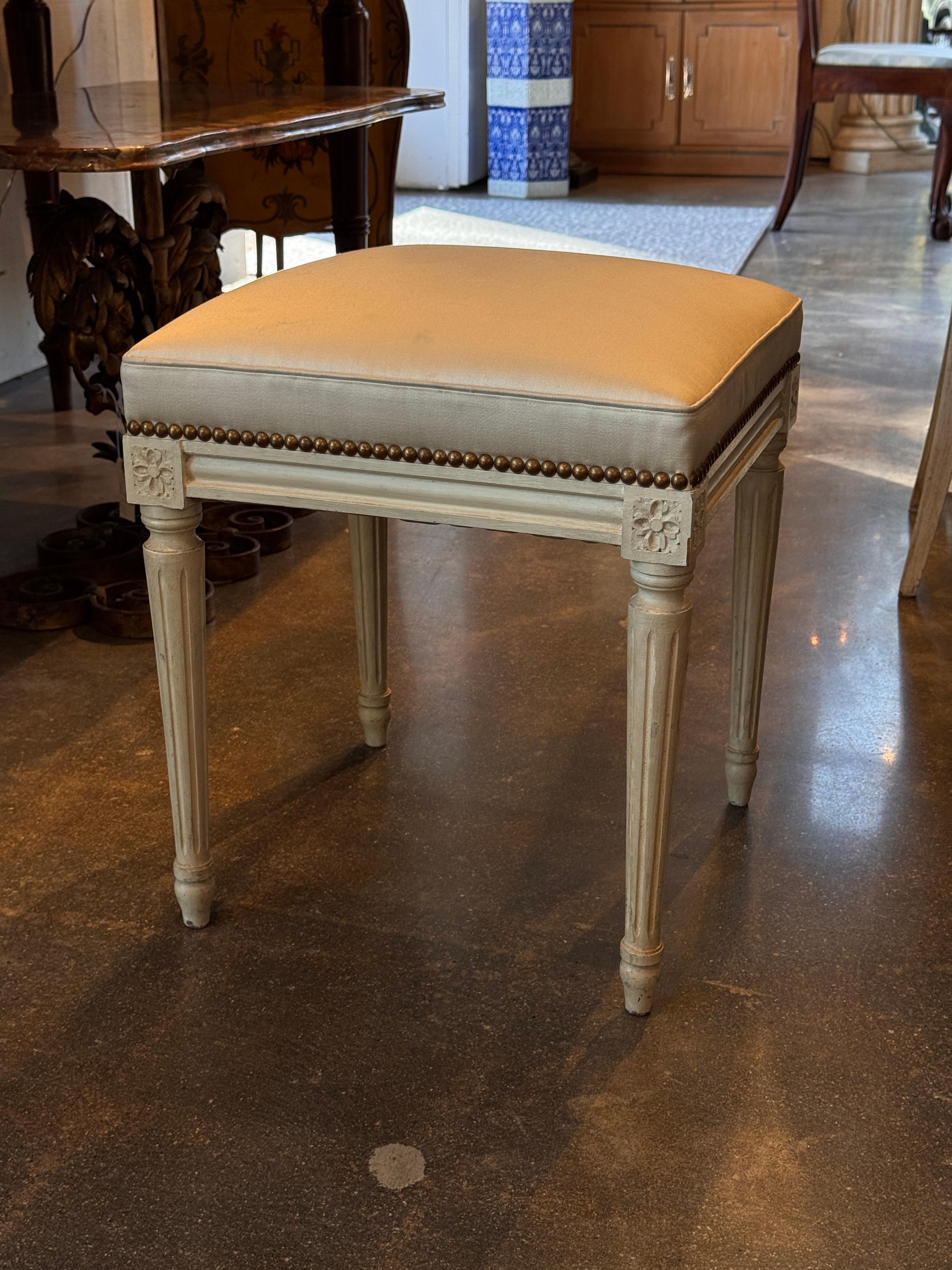 Bemalter Hocker im Louis-XVI-Stil mit kannelierten, spitz zulaufenden Beinen, geschnitzten floralen Eckblöcken und Messingnägeln entlang des gepolsterten Sitzes. Die Nahaufnahme zeigt die scharfen Schnitzereien und die originale Lackierung.