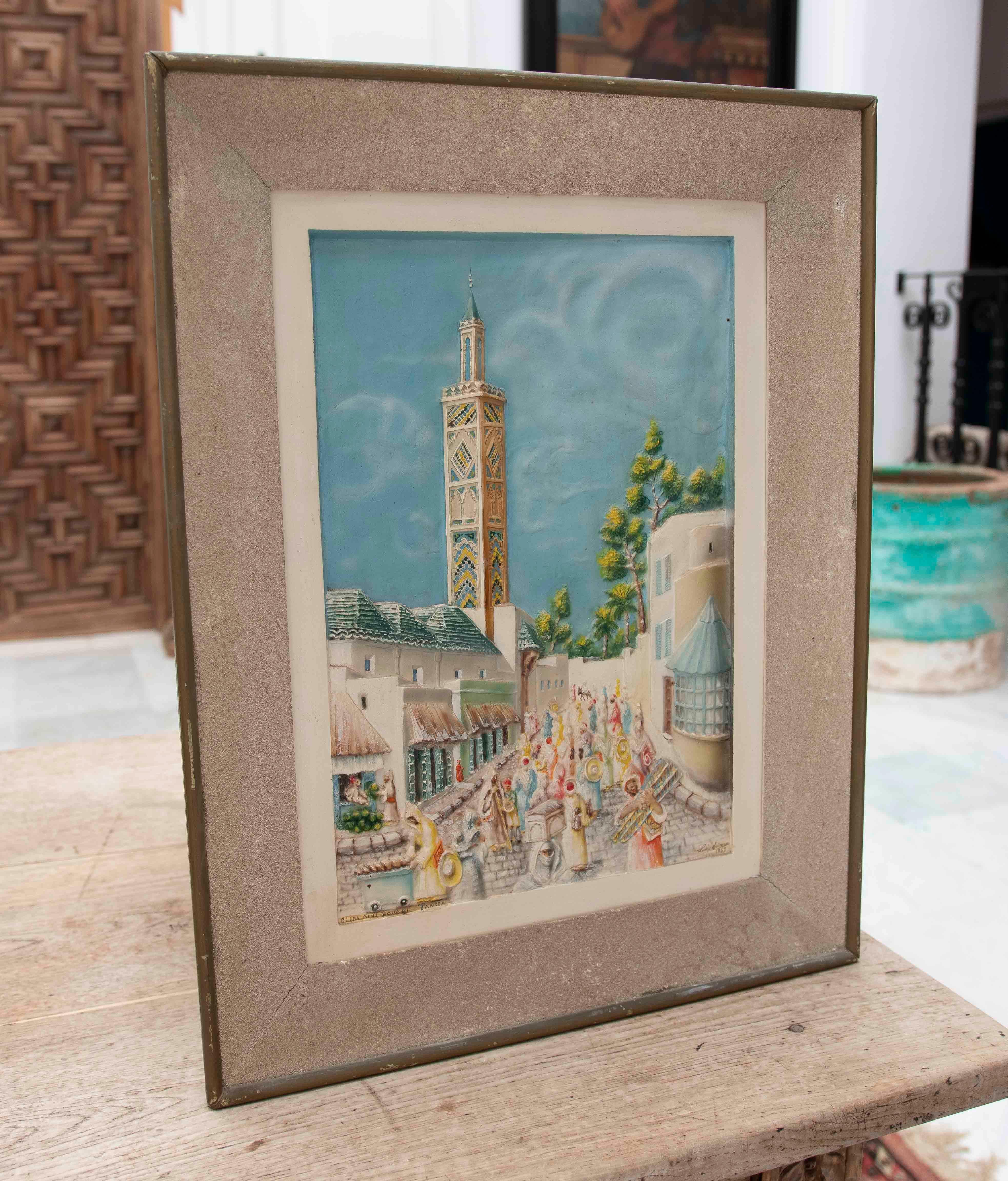 Pintura en relieve sobre papel que representa una animada escena urbana con arquitectura tradicional norteafricana. Un ornamentado minarete destaca en el centro, rodeado de edificios con tejados inclinados y paredes blancas. El primer plano muestra