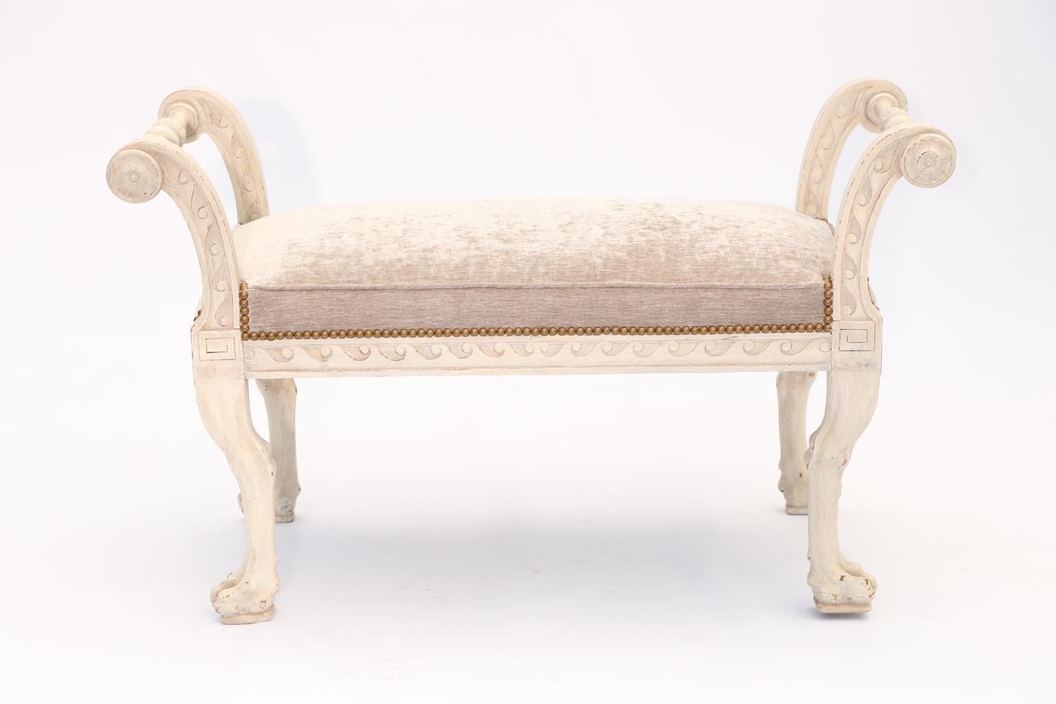 Bench, in Regency style, having a painted finish showing natural wear, its apron carved with evolute scroll, extending to its outswept arms, joined by turned stretcher, decorated with rosettes, flanking a crown seat of velvet, upholstered with