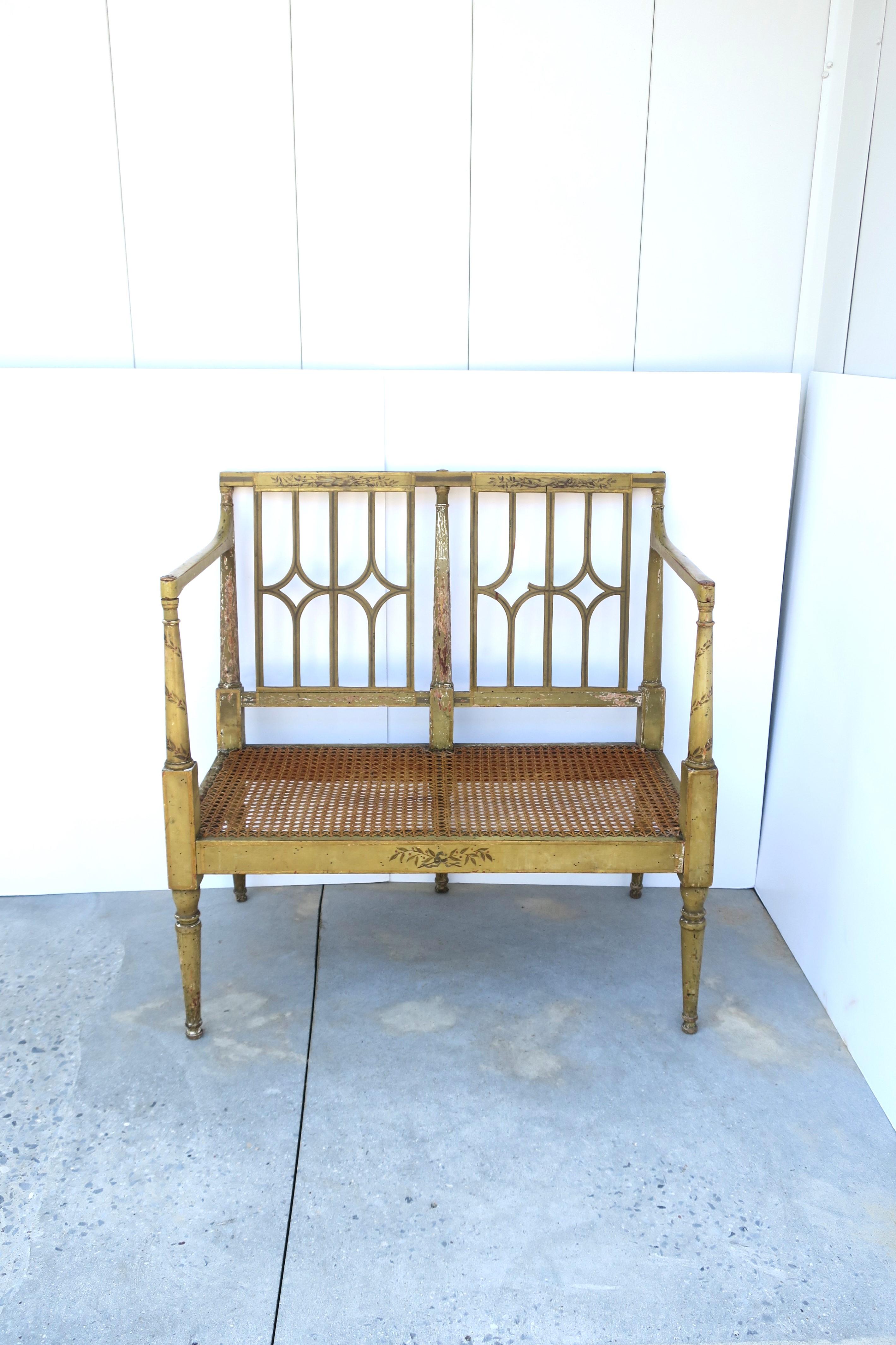 A painted and distressed wood and cane wicker loveseat bench, circa mid-20th century. Piece has a perforated back, high arms, cane seat, decorated accents, finished with an intentional distressed look. Seat height: 13.75