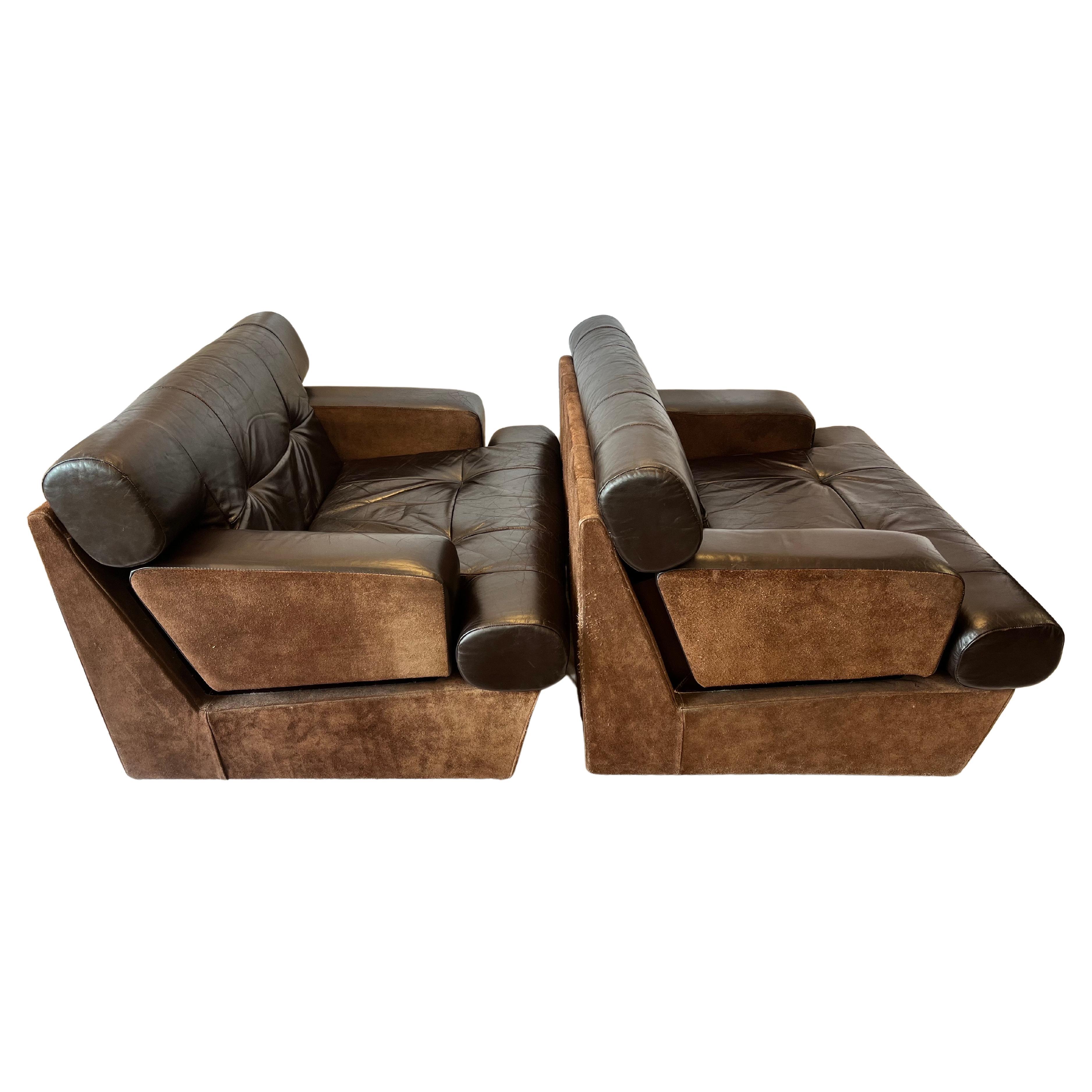 A pair of circa 1980's era leather and suede lounge chairs presented in a deep chocolate brown hue and accented by wood design elements visible when experiencing the lounge chairs from the side. The low and confident design is wide and expansive