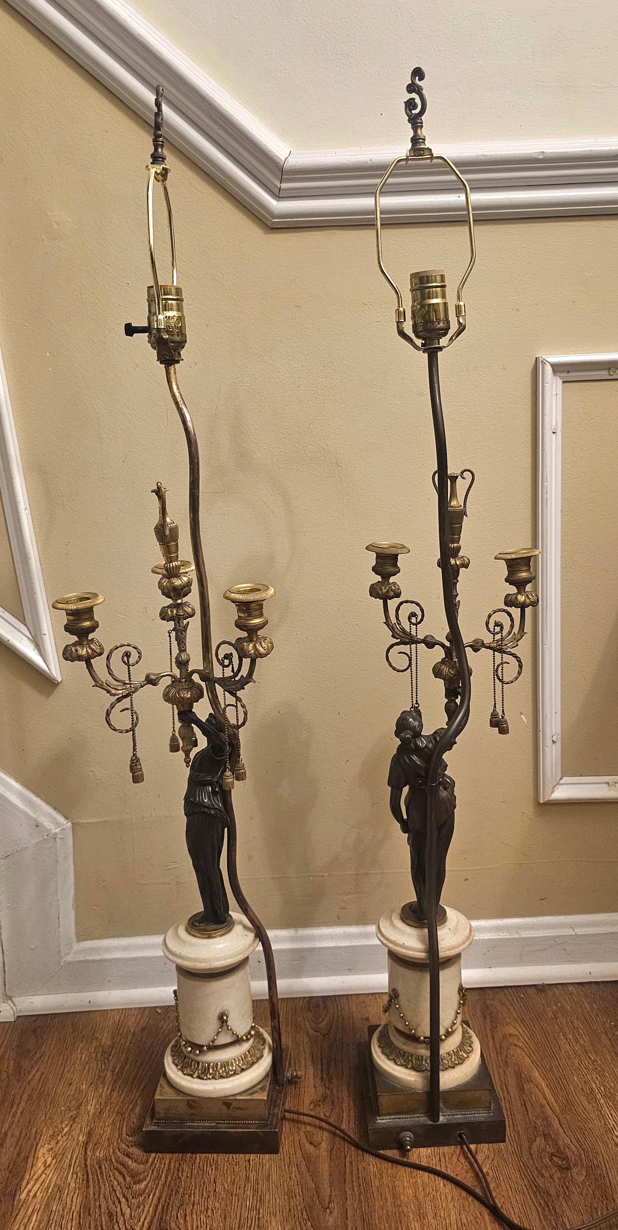 
Coppia di candelabri figurativi francesi in bronzo Impero del XIX secolo su basi a plinto in marmo di carrara, ciascuno con bordature e finiture in bronzo dorato,  ora montate come lampade, ciascuna modellata come una fanciulla che regge tre