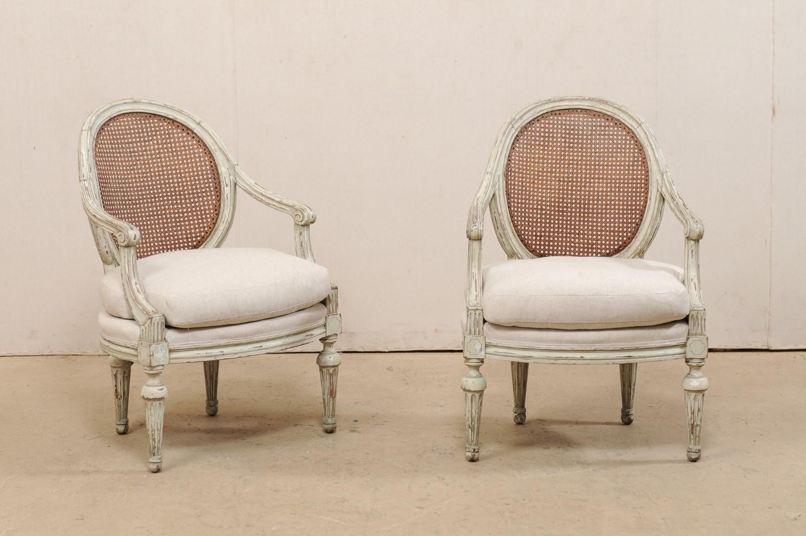 A French pair of Louis XVI style round cane-back and upholstered seat armchairs from the 19th century. This pair of bergères chairs from France each features a rounded back, handwoven with cane, beautifully scrolled knuckles at the end of