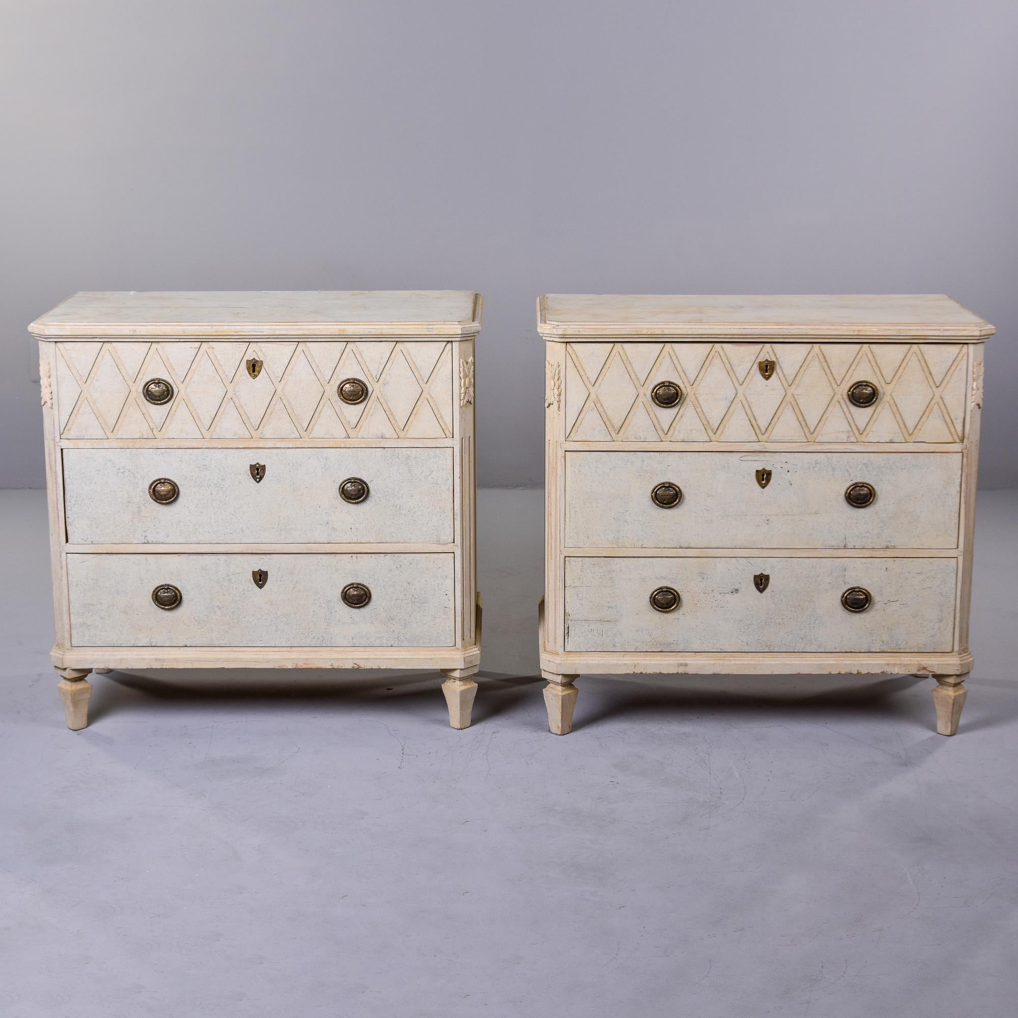 Pair of circa 1880s Swedish three drawer chests with bone colored painted finish. These chests have carved diamond pattern on top drawer fronts, dovetail construction brass pulls and escutcheons and carved medallion details toward the top on the