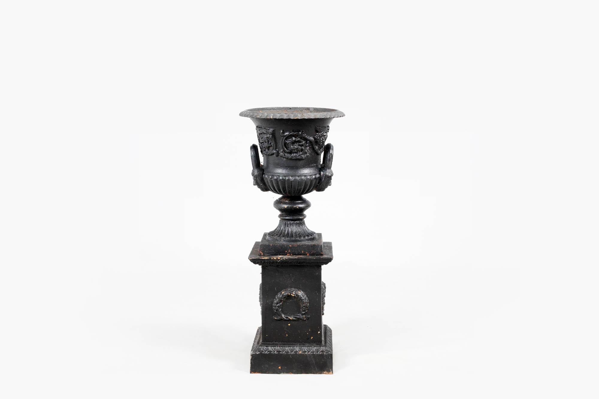 Pair 19th century Regency era cast iron Campana-shaped petite garden urns. The pair feature Classical style design with dentil detailed rims, decorative foliate relief to the bodies, and flared masked handles on reeded round bases. Each urn sits on