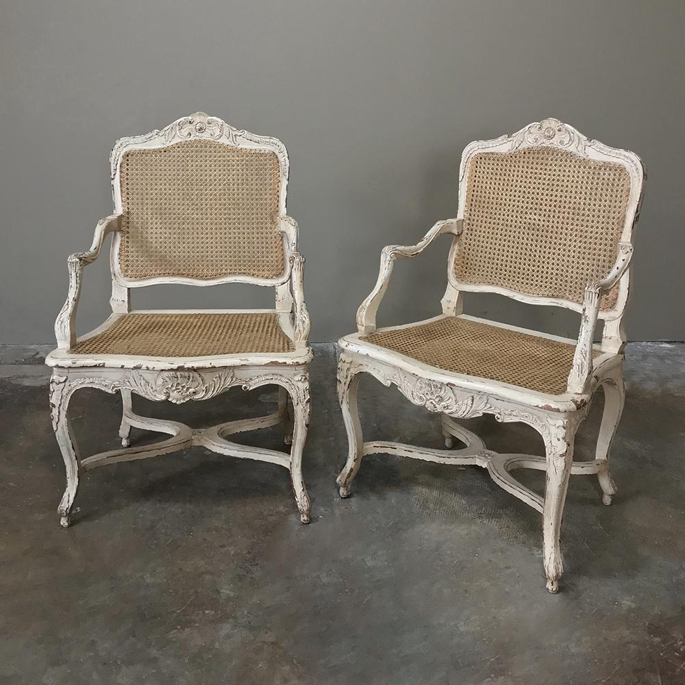 Pair of 19th century Country French caned painted armchairs are magnificently carved from the arched seatback crown to the graceful cabriole legs below, and the painted finish has achieved a level of attractive distress that is so in vogue today.