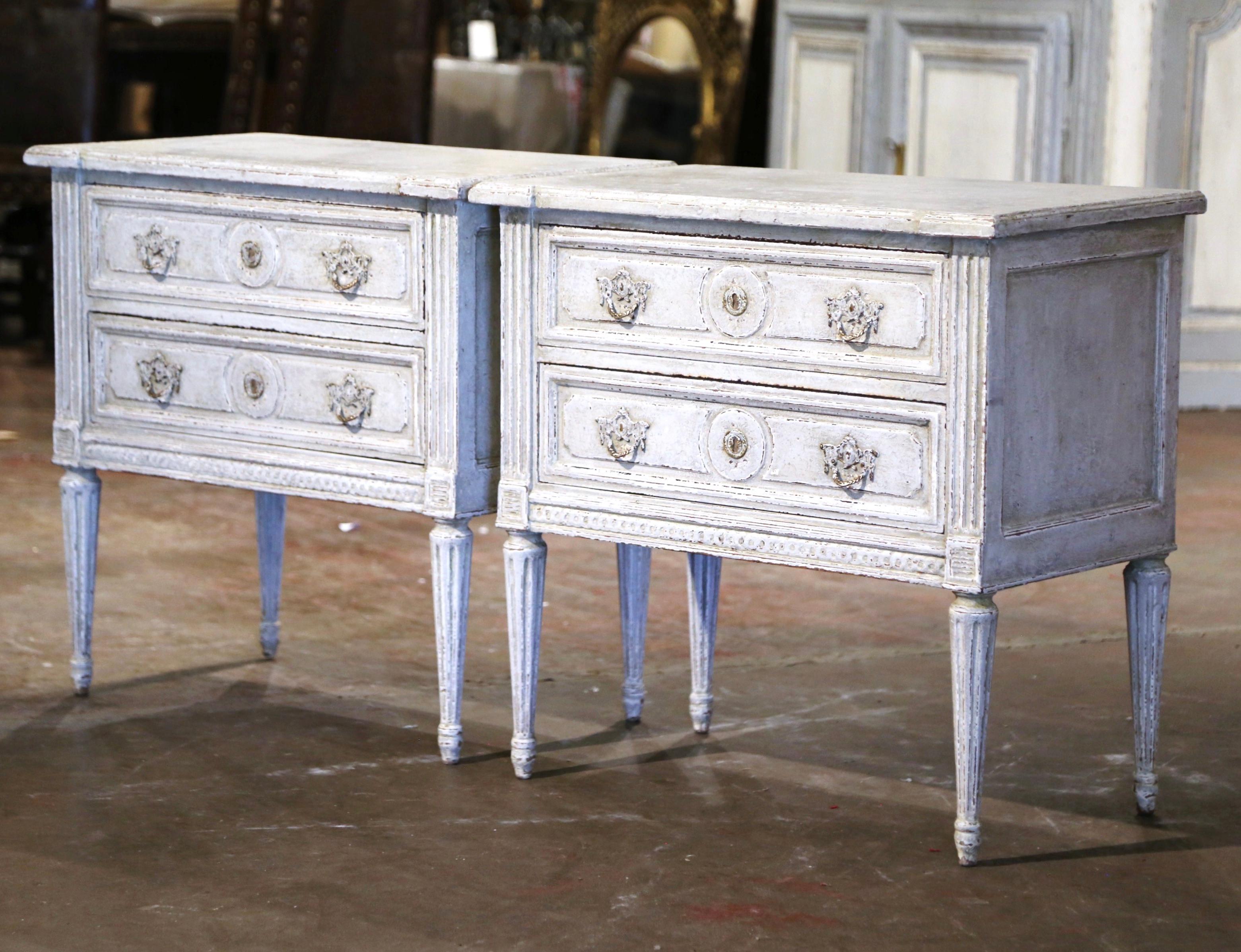 Complete your bedroom with this elegant pair of antique painted bedside cabinets! Crafted in France, circa 1880, each commode stands on tall tapered fluted legs decorated with floral rosette medallions at the shoulder. The chest with side cut spline