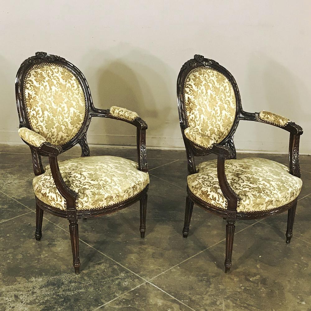 Pair of 19th century French Louis XVI walnut armchairs feature contoured oval seatbacks and generous seats with padded armrests for surprising comfort! Hand carved along the entire solid walnut framework, each is upholstered in damask fabric that is