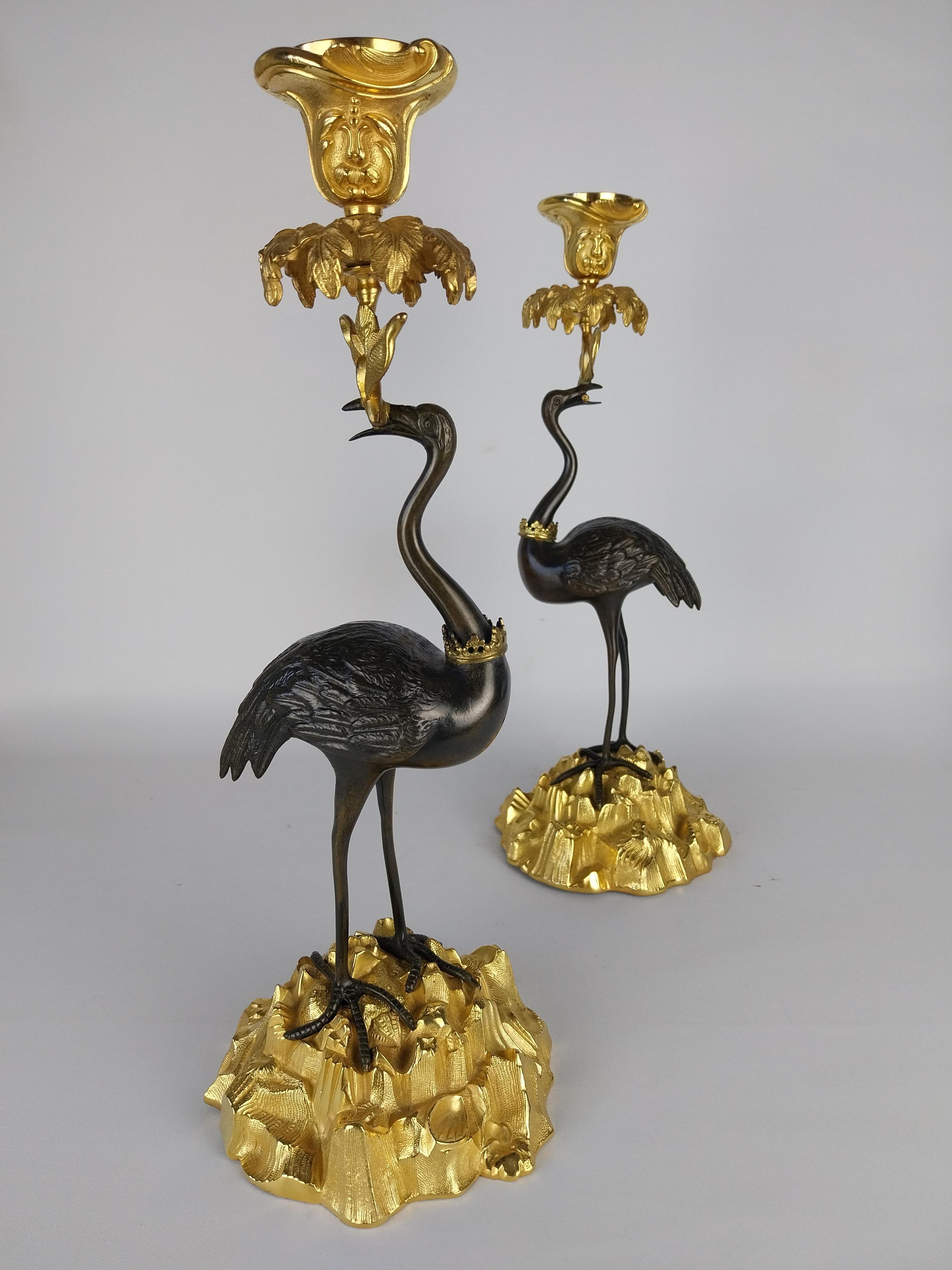 Coppia di candelieri del XIX secolo fusi a forma di cicogne in bronzo, ciascuna con una corona dorata intorno al collo e che tengono una singola candela nel becco, in equilibrio su basi di roccia e conchiglie naturalistiche dorate, appesantite da un