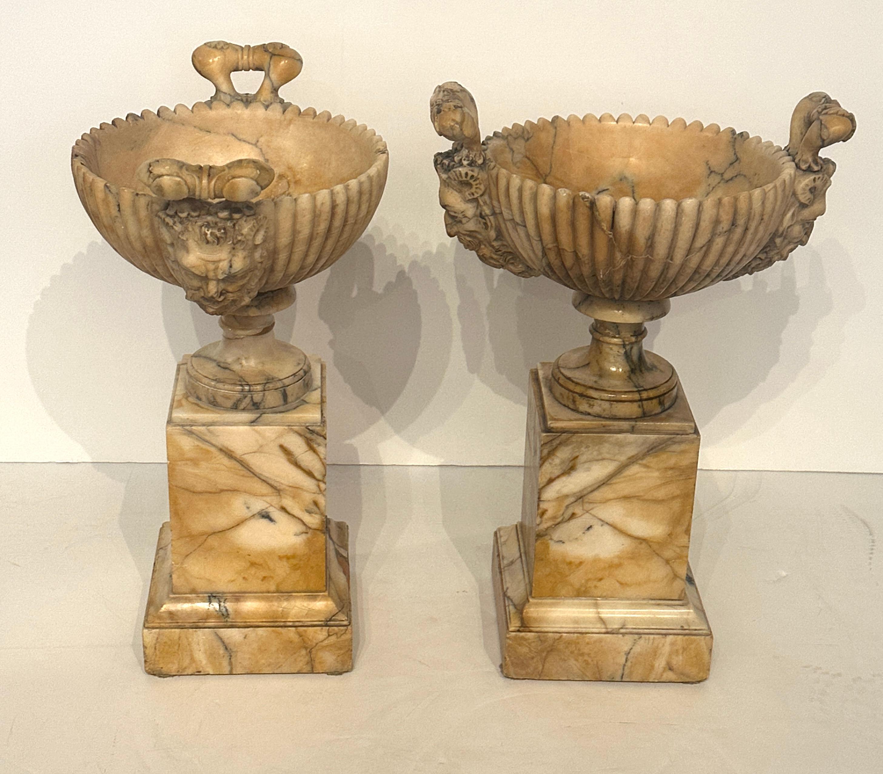 A hand-carved pair of alabaster coupes with attached stepped pedestals. They feature figural handles and a center relief of a flower.
