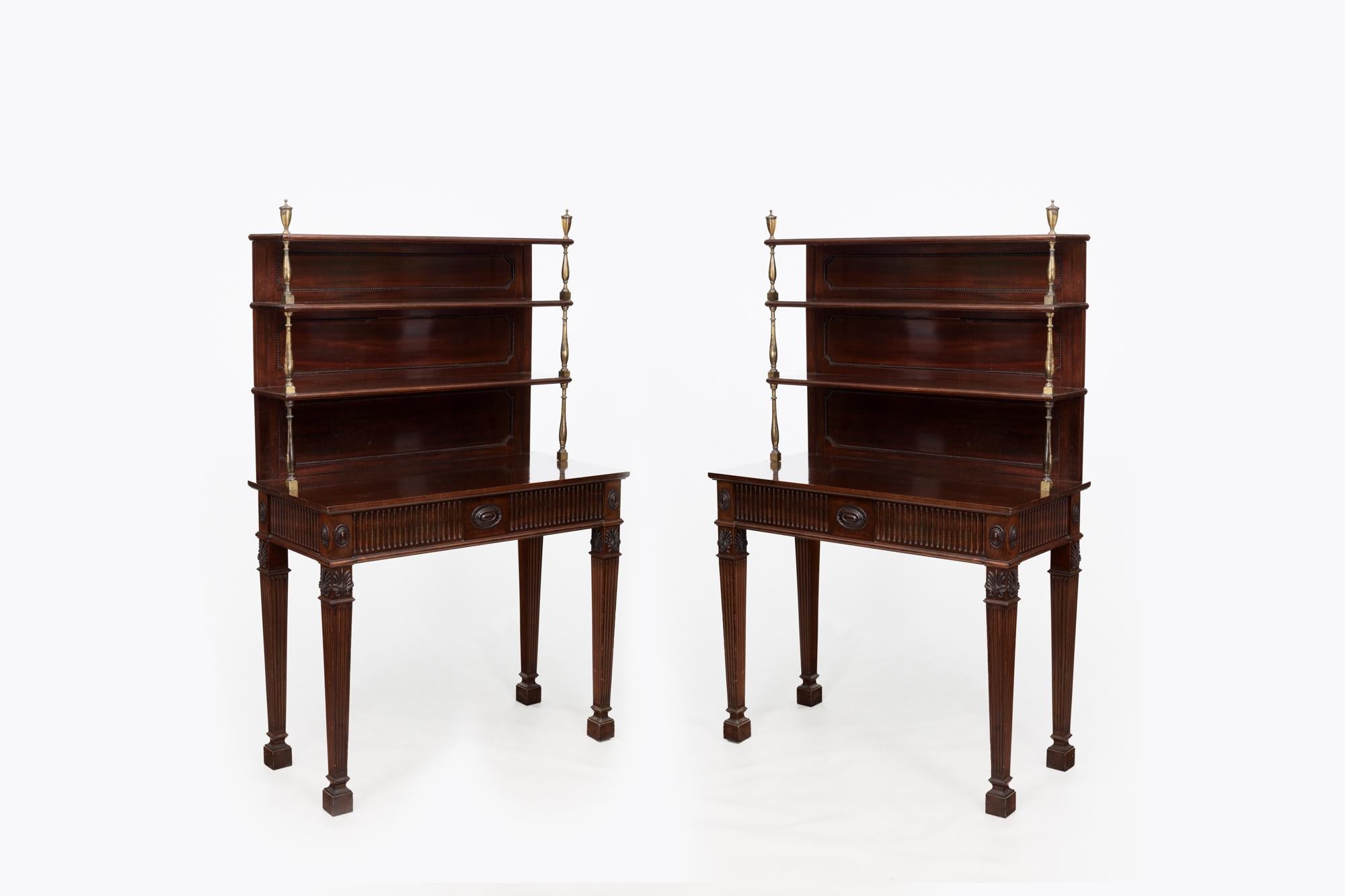 Pair 19th Century Hepplewhite mahogany chiffoniers with open shelving to the upper section supported by brass uprights topped with classical urn-shaped finials. The lower sections, in the Adam style, feature carved oval paterae and fluted details to