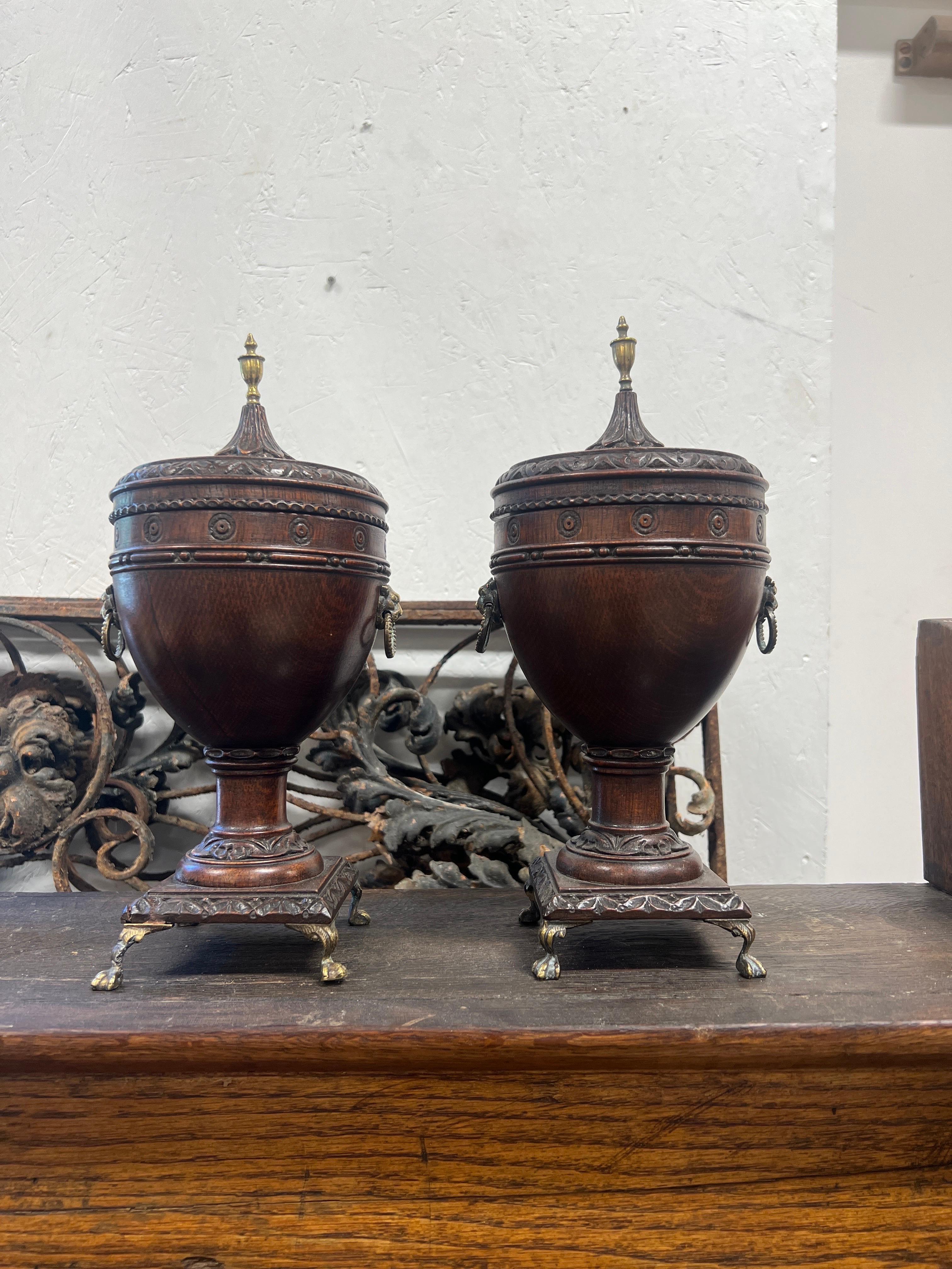 English, early to mid 19th century.

A fantastic pair of Regency urns. Each having a oak body with foliate carved leaf trim to borders, bullseye border to neck and good quality brass lion mount pulls, paw feet and federal style finial mount.
