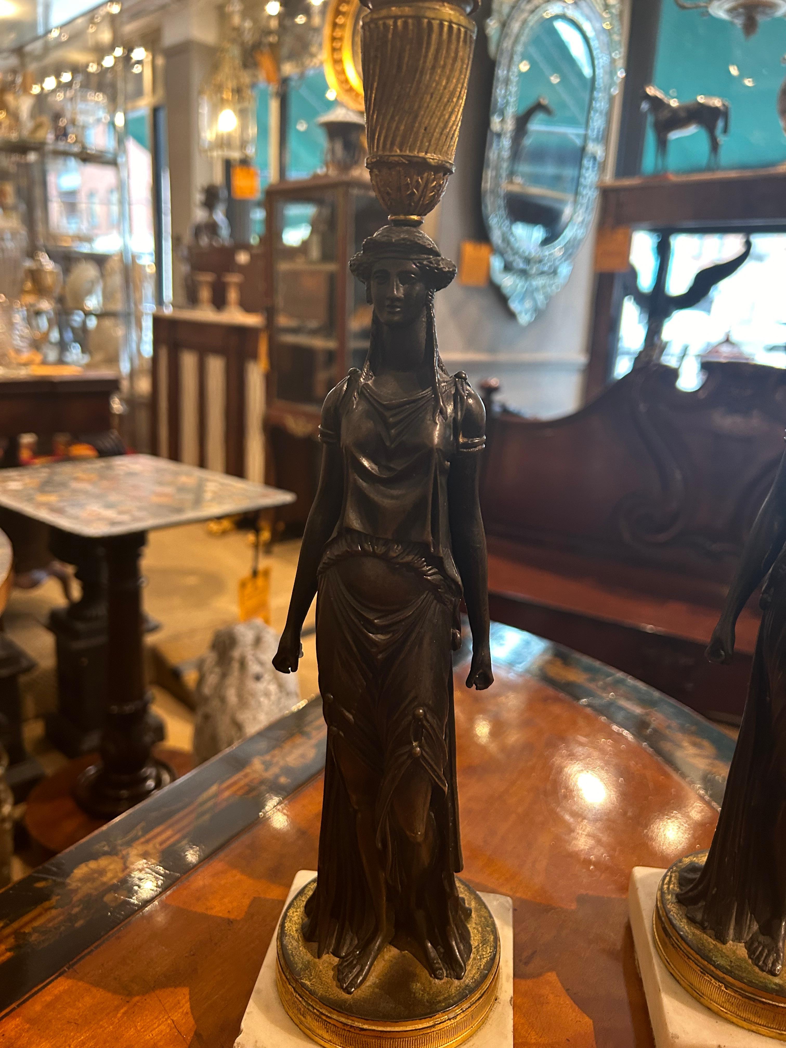 Pair 19th Century Regency bronze candlesticks with ormolu detail in the manner of William Kent. The pair depict two female figures in Grecian dress, likely water carriers. They sit on white squared marble bases. Circa 1810.