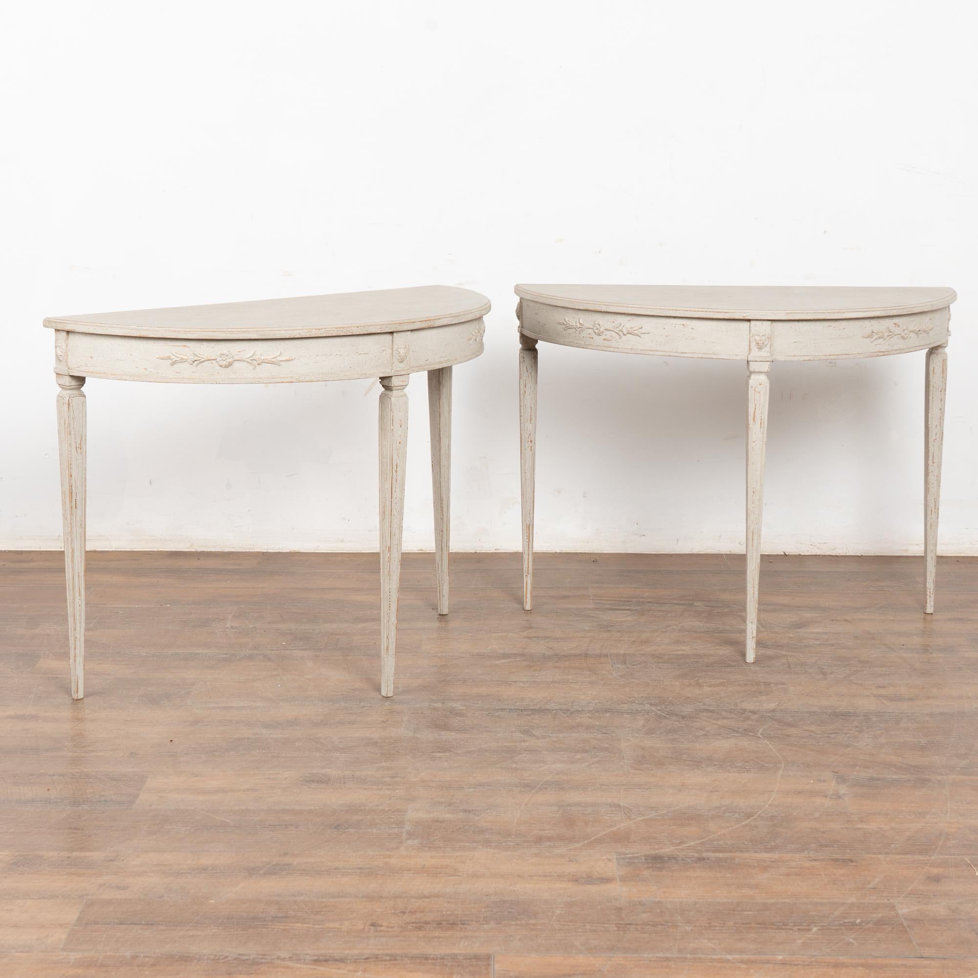 Pair, Gustavian style demi-lune side tables or consoles. 
Gently tapered long fluted legs topped with decorative applied lion's head carving and floral swag along skirt.
Newer professionally applied layered antique white painted finish with gray
