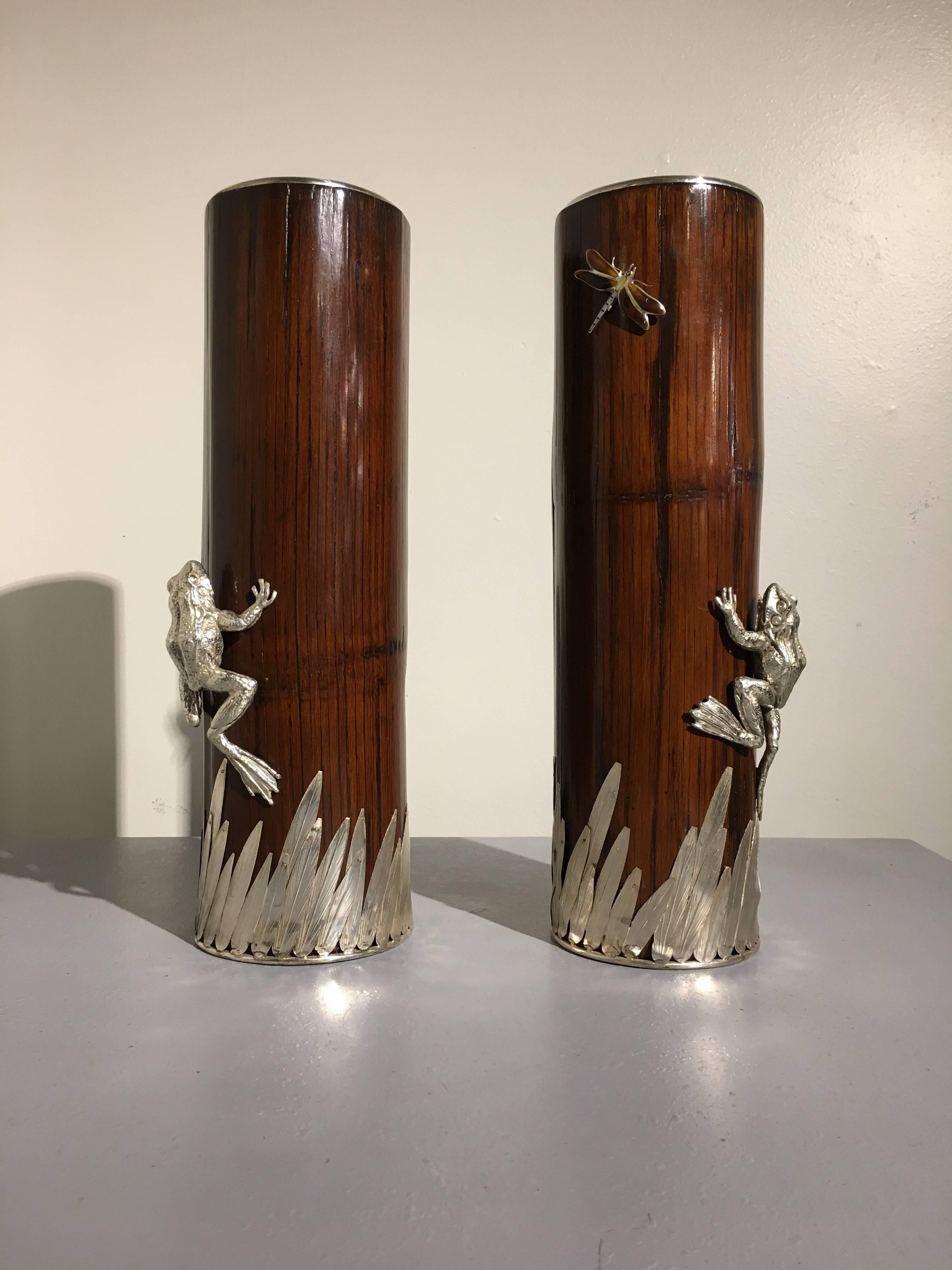 A large pair of silver plate mounted bamboo vases. The tall vases of cylindrical form each feature a natural section of dark brown bamboo, with leaves of silver plate, as well as a large silver plate frog and a dragonfly.
The frog is portrayed