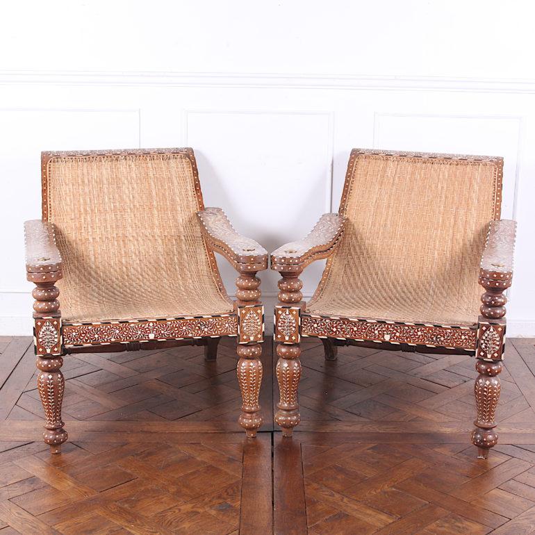 A very unusual pair of profusely-inlaid plantation chairs, the teak frames entirely covered with bone and ebony inlay. The caning has been replaced and is in very good condition. These low-slung lounge chairs feature swing-out arms for kicking ones