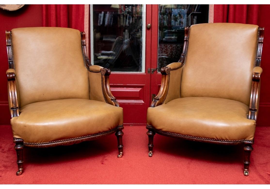 A pair of Classic English style club chairs. The elegant chairs with closed arms, the carved mahogany frames with scrolled side and arm supports. Upholstered in a tan leather with nail head trim. Raised on turned front legs on casters and curved