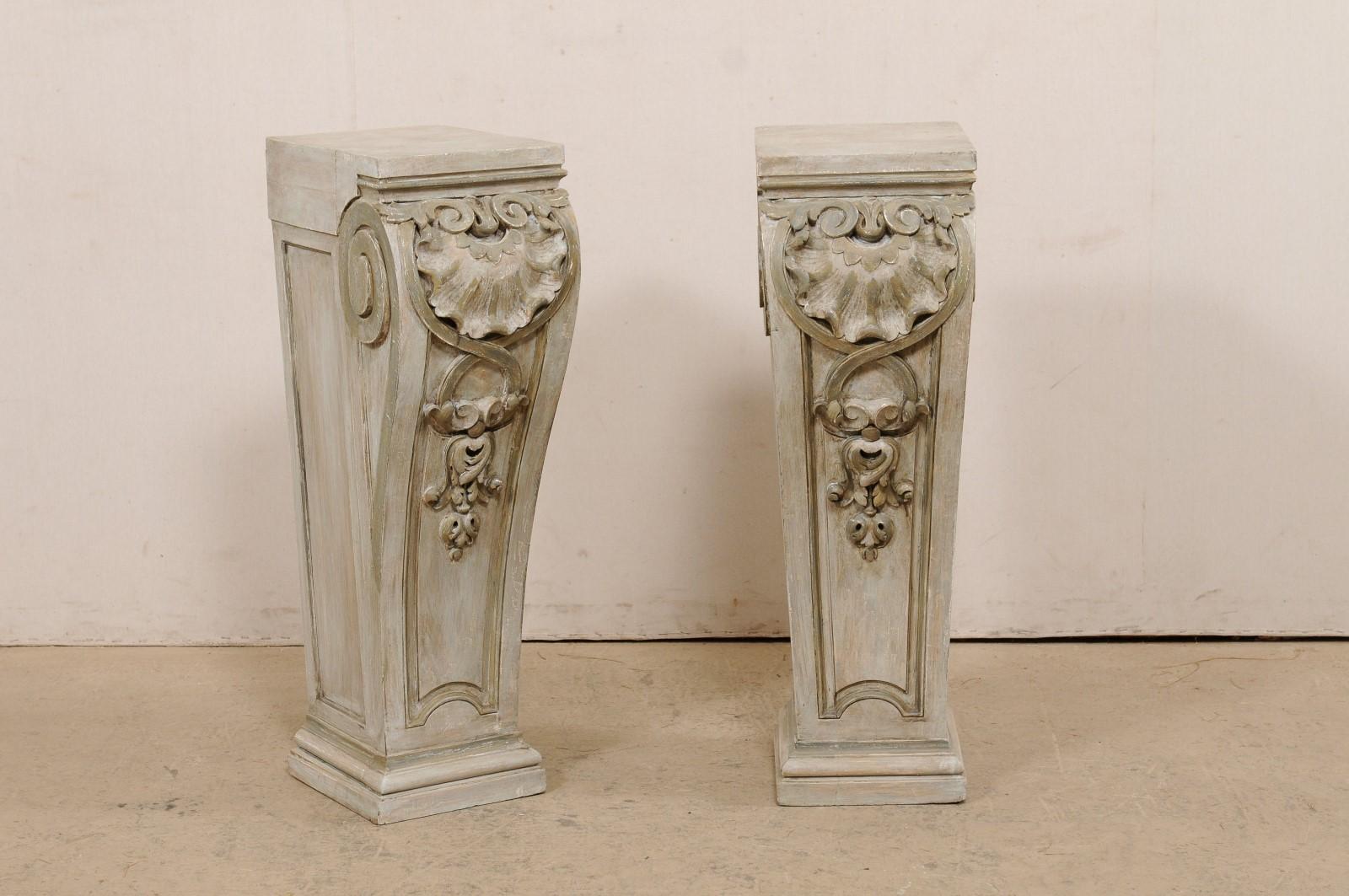 A pair of American carved-wood pedestals from the early 20th century. This antique pair of pedestals have been carved in a shell, foliage and volute motif. The rectangular-shaped tops are flat, and stick out farther from the lower more squared