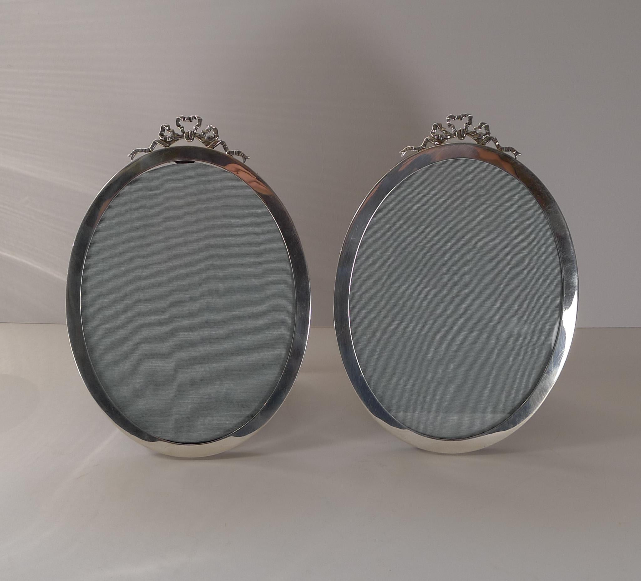 A stunning pair of oval photograph frames, made from English sterling silver topped with ribbon and bow mounts.

The backs are made from solid oak incorporating a folding easel back stand.

The silver is fully hallmarked for Birmingham 1915