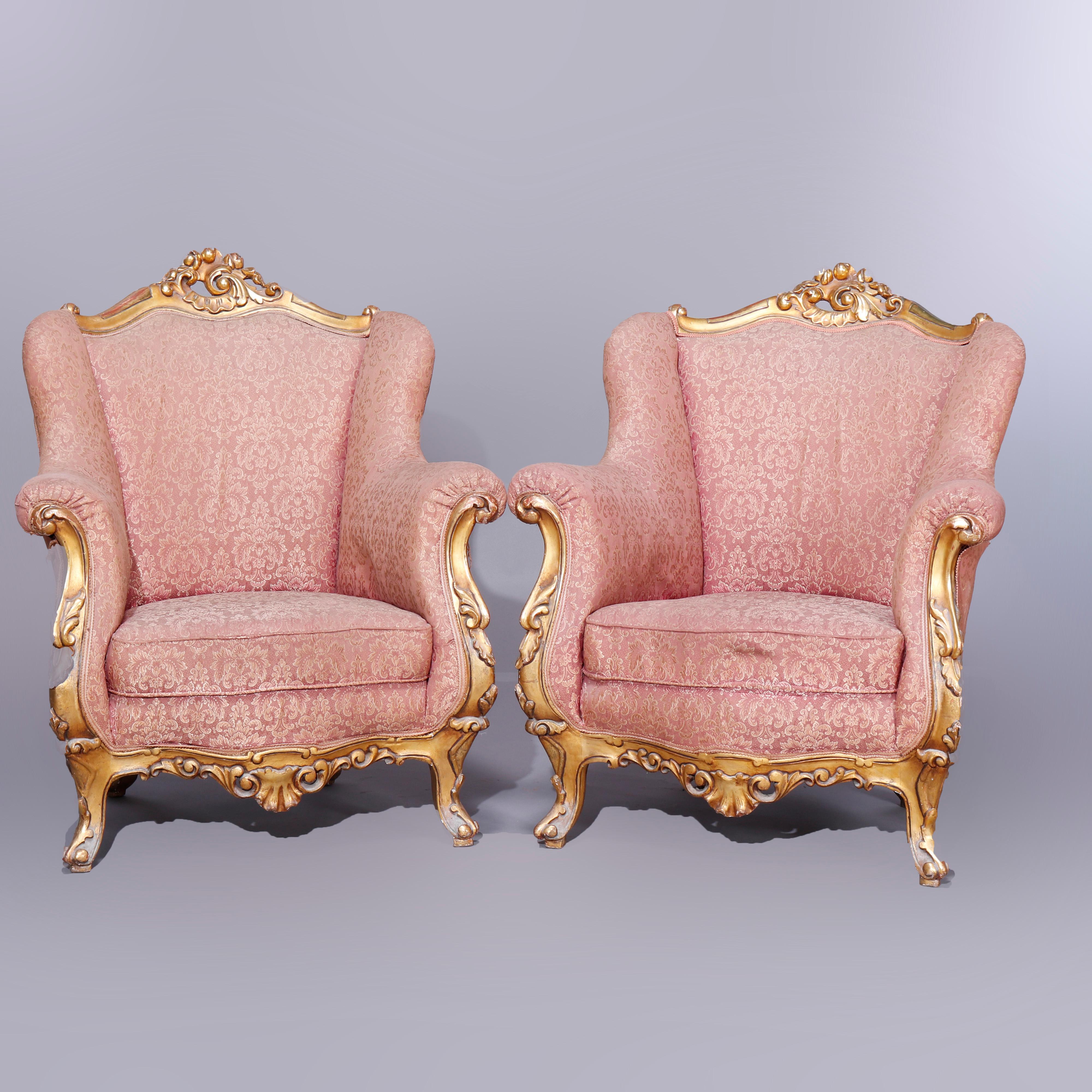 A pair of antique French Louis XIV style wing back bergere armchairs offer pierced and foliate carved gilt crest over upholstered wing back fireside chairs having scroll form arms and foliate carved giltwood apron, raised on giltwood cabriole legs,