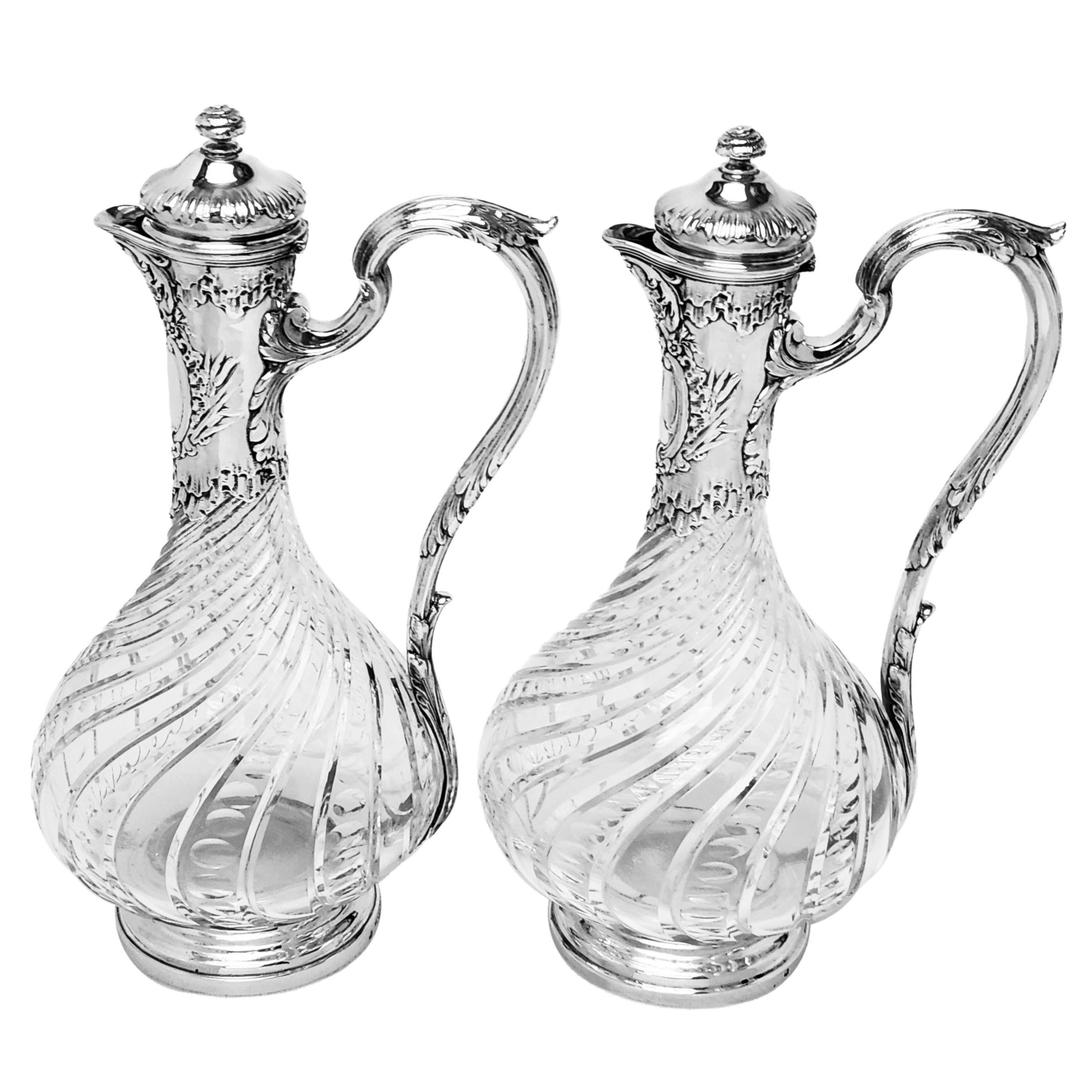 A pair of elegant Antique Silver mounted Glass bodied Claret Jugs made in Paris, France. This pair of Wine Jugs has an ornate chased and engraved Neck and lid with a shaped cartouche below the spout. The Body of each Jug is decorated with a writhen