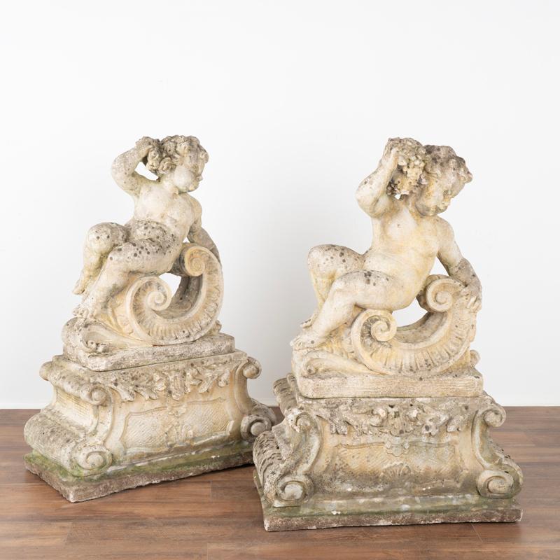Classical garden statue of putti representing two seasons. The stone composite statue figures each depict a cherub with textured curly hair, rounded cheeks and naked body with one holding roses and the other a cluster of grapes, both reclined on a C