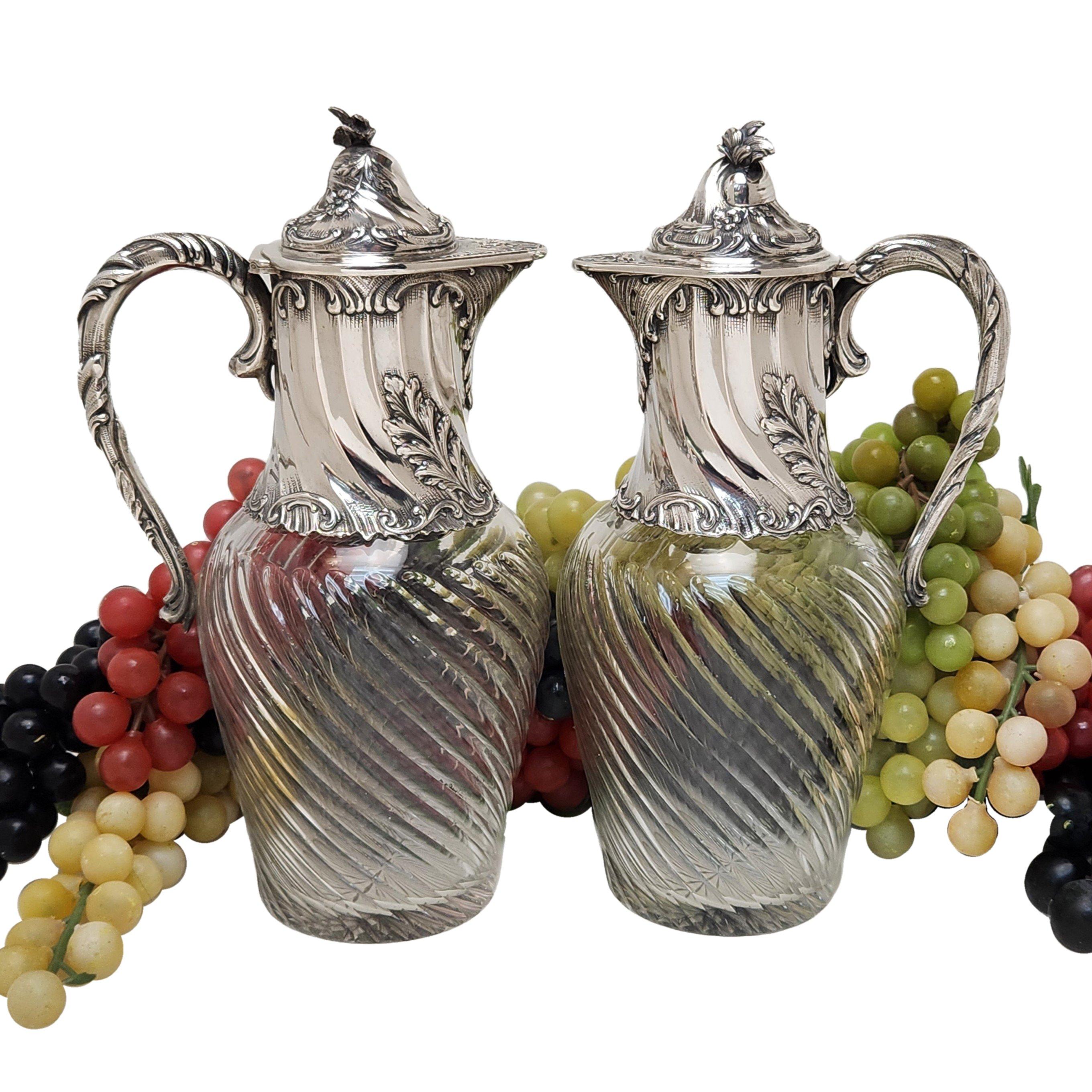 A pair of elegant solid Silver & Glass Claret Jugs. Both the Glass Bodies and Silver Necks of the Jugs are embellished with a writhen fluted pattern. The neck, hinged lid and scroll handle of each jug feature lovely chased and engraved scroll and