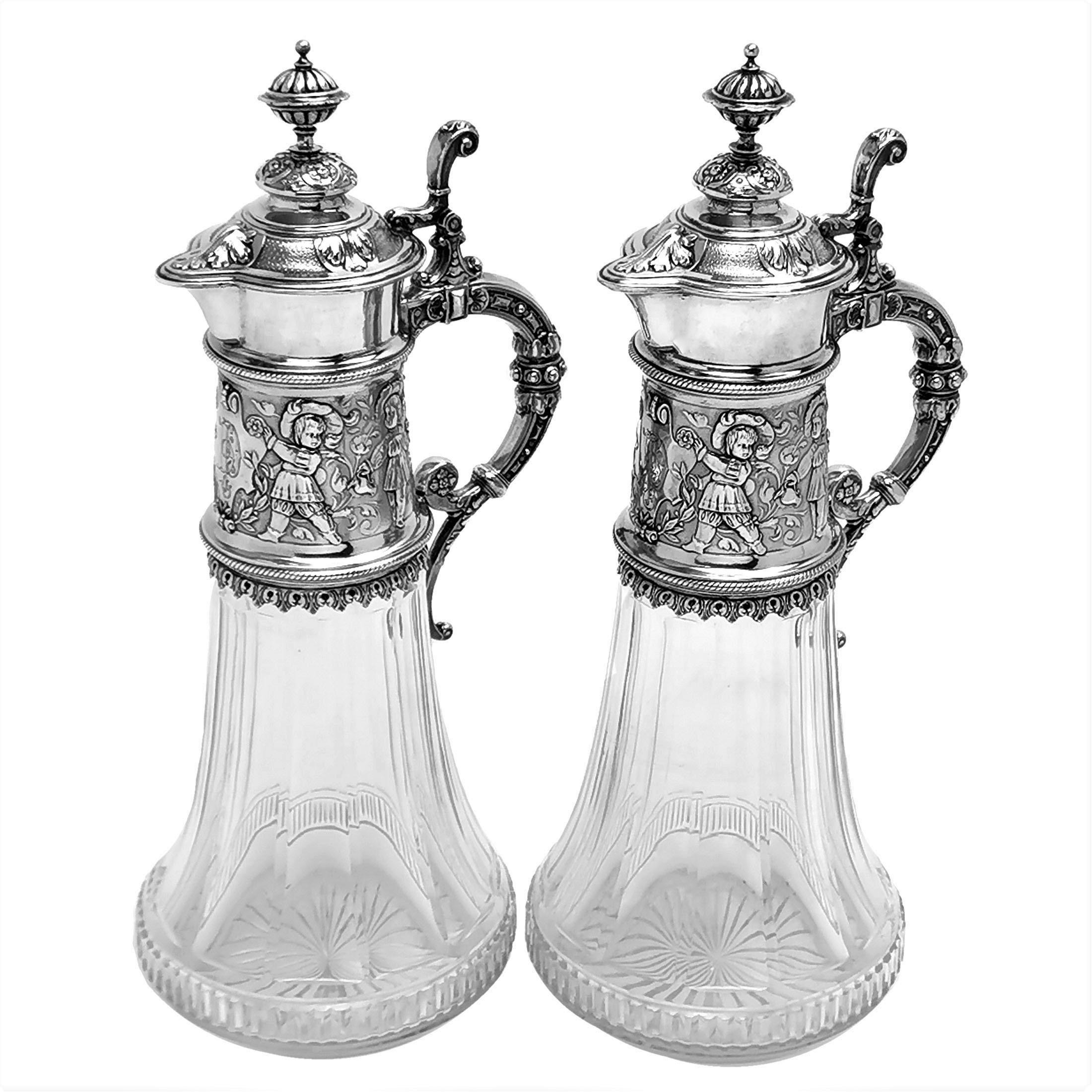 A pair of beautiful Antique German silver & glass Claret Jugs with an elegant fluted cut glass body and an ornate solid Silver neck, handle and hinged lid. The wide neck of the Wine Jug has a band of intricately chased decorations showing a pair of