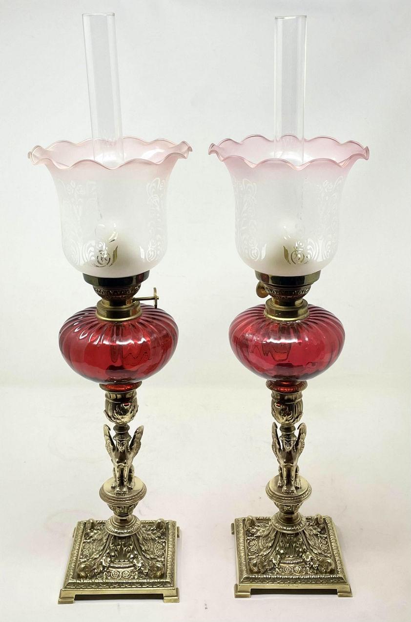 A fine pair of stylish and imposing French grand tour style polished brass table oil lamps of compact proportions modelled to depict two identical seated Winged Sphinx below a Cranberry glass reservoir, clear glass chimney and vaseline frosted glass