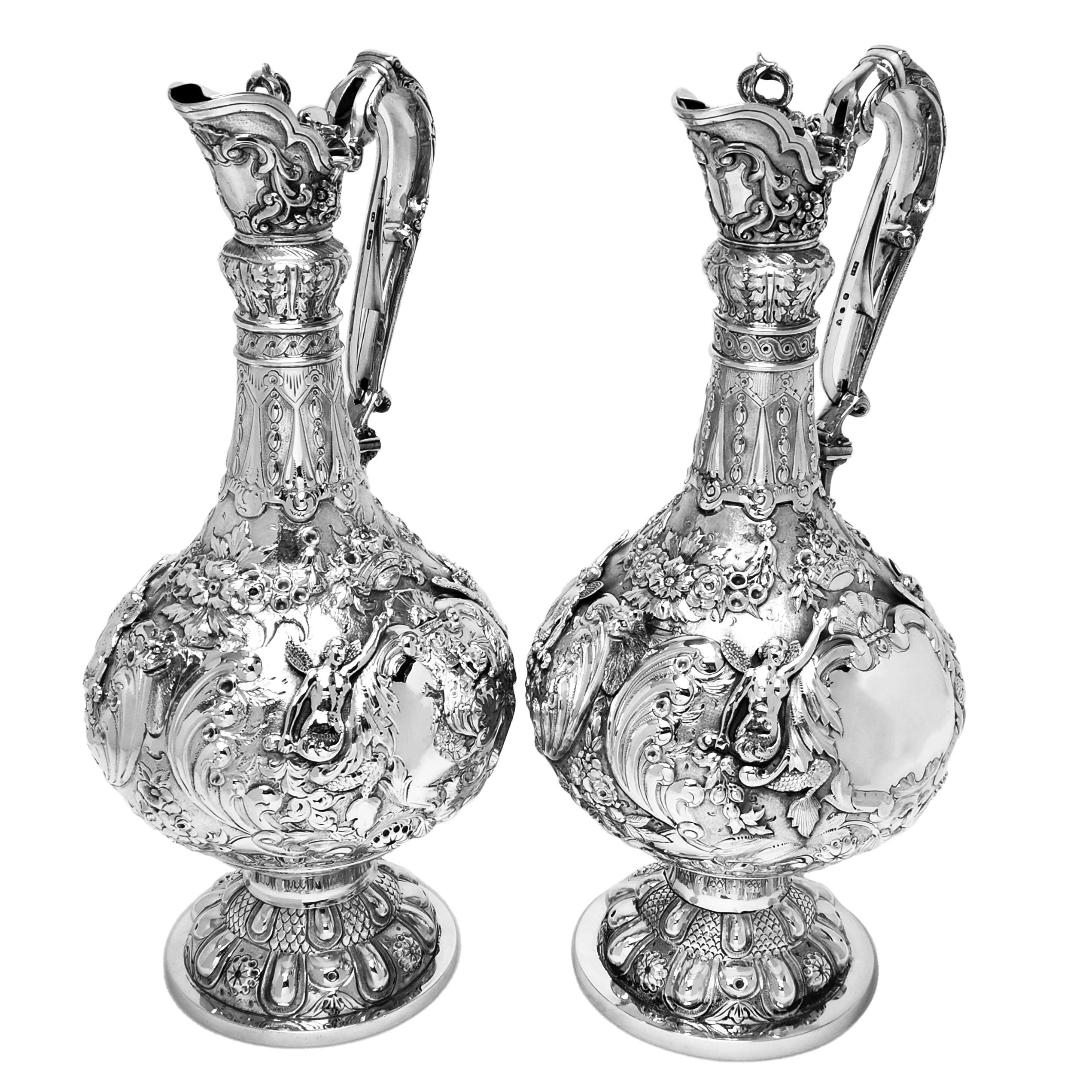 An impressive pair of Antique Irish sterling silver Claret jugs in a traditional Armada Style. This matched pair of Armada Wine Ewers feature rich and ornate chased and applied detailing over the entire body of the jug. These chased decorative