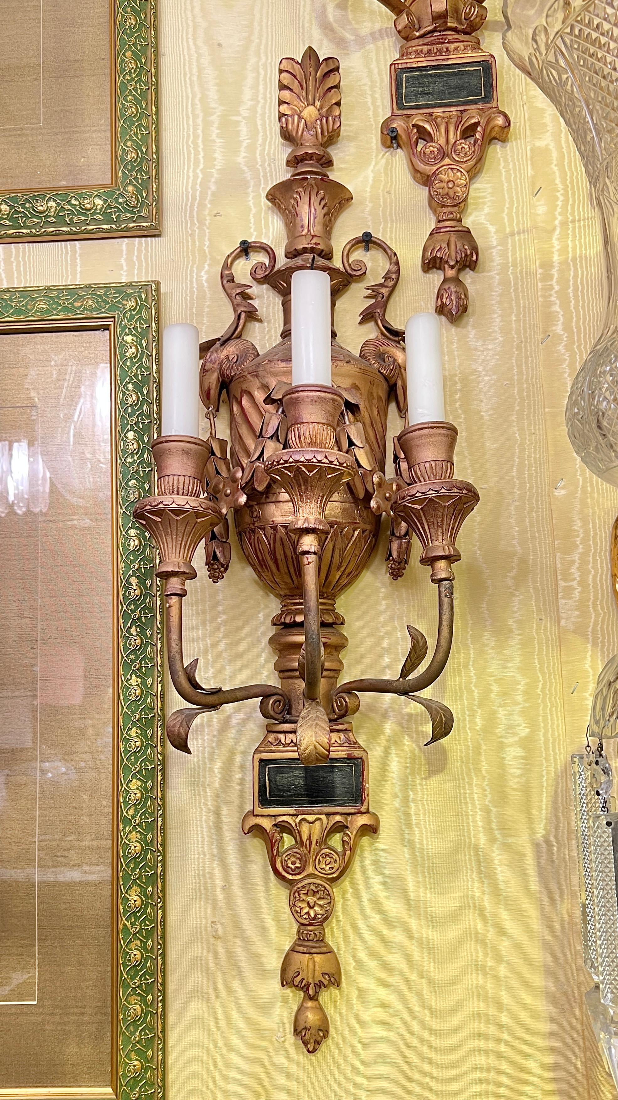 Pair of antique three-light sconces in the neoclassical style crated from wood and tole metal with gold finish, of urn form with stylized ram handles, floral swags, feathers and foliate scrolls.  Each 30 by 9 by 7 1/2 inches with wax candles.