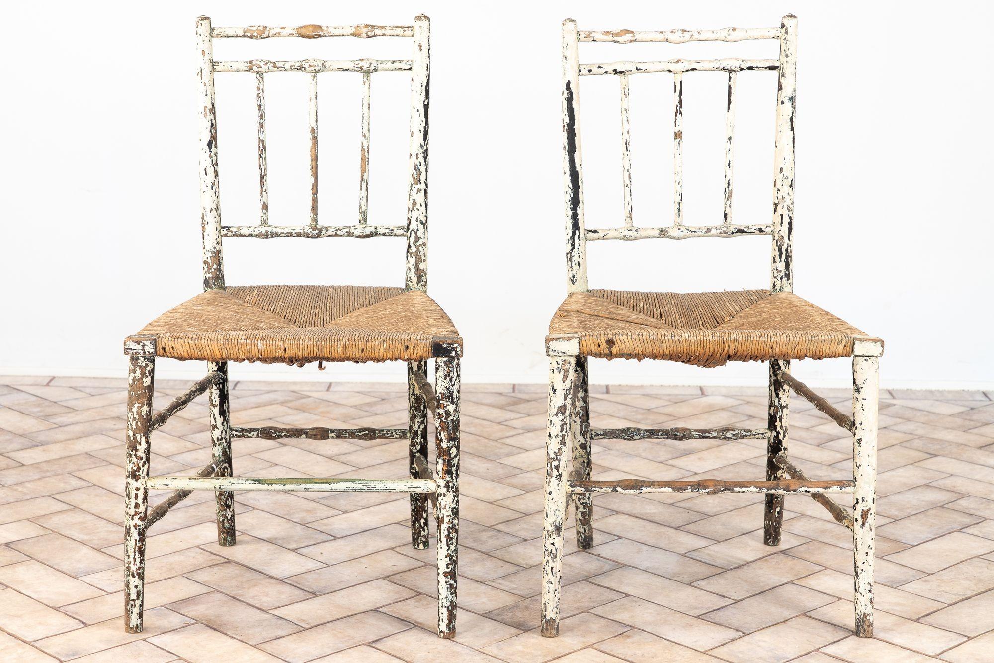 Une charmante paire de chaises rustiques de ferme anglaise peintes avec des sièges en jonc tissé à la main et une peinture blanche en couches magnifiquement usée. Les dossiers simples en échelle et les détails tournés offrent un style campagnard